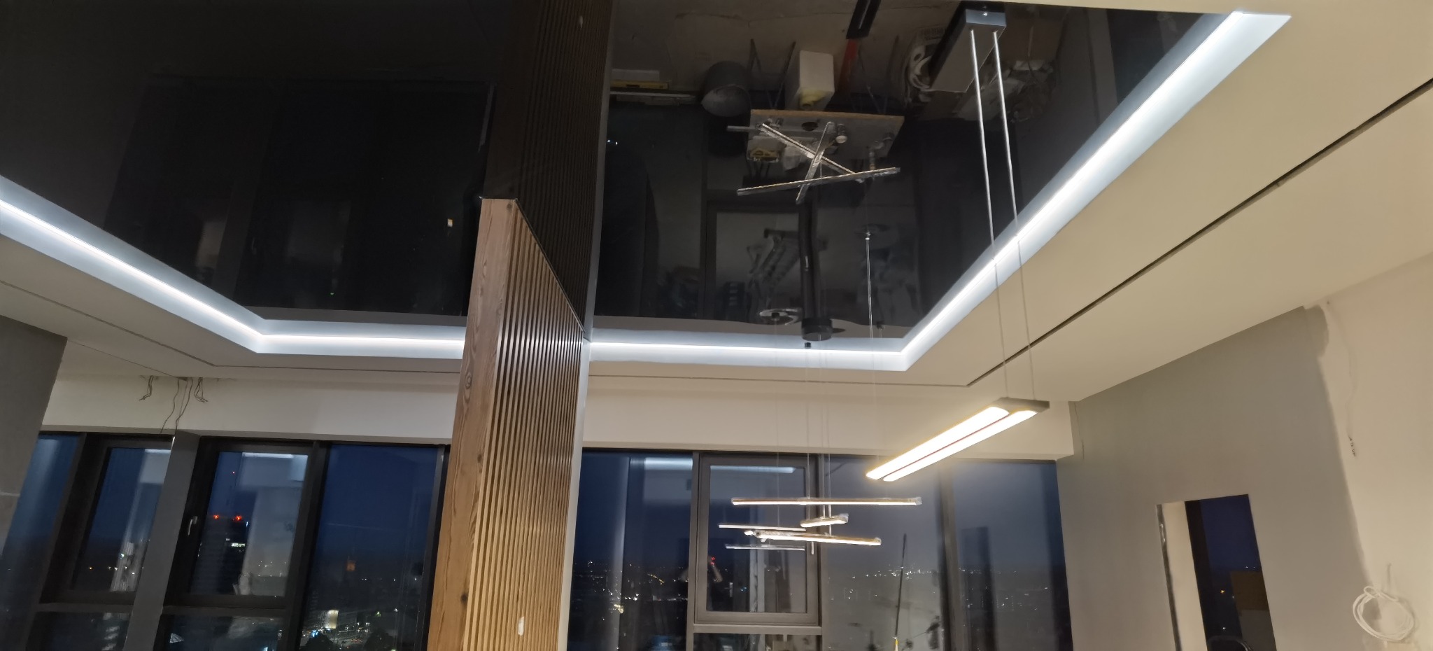Czarne lustrzane wykończenie sufitu z oświetleniem LED, odbijające lampę i przestrzeń roboczą, widoczne duże okna z widokiem na miasto.