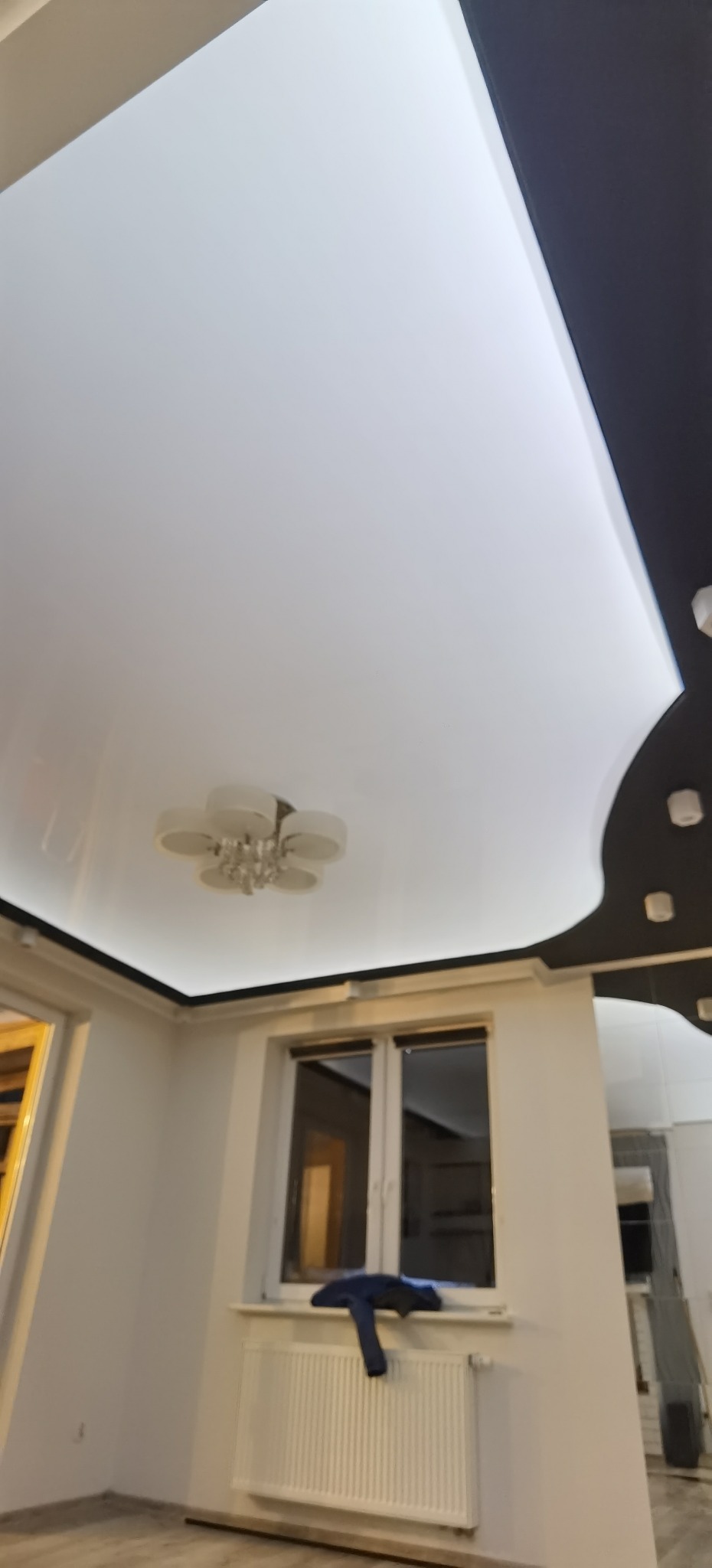 Biały, błyszczący sufit z czarnym obramowaniem w kształcie fali, oświetlenie LED ukryte w obramowaniu, nowoczesny żyrandol w kształcie kwiatu, okno z niebieską kurtką na parapecie.