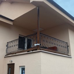 TECH-WELD Wojciech Putrym - Czarna, metalowa barierka balkonowa z ozdobnymi elementami, zamontowana na balkonie domu o beżowej elewacji. Widoczna kamera monitoringu przymocowana do barierki.