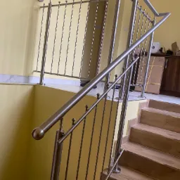 Wewnętrzna klatka schodowa z drewnianymi stopniami i nowoczesną, metalową barierką z pionowymi prętami, zakończonymi kulkami, na tle żółtej ściany.