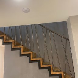 TECH-WELD Wojciech Putrym - Nowoczesne schody z drewnianymi stopniami i metalową balustradą o minimalistycznym designie, widok z dołu na szarej ścianie, z taśmą przyklejoną do poręczy.