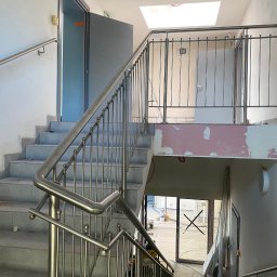 TECH-WELD Wojciech Putrym - Klatka schodowa w trakcie wykończenia, widoczne schody wyłożone szarymi płytkami, metalowe barierki z pionowymi prętami i okrągłymi zakończeniami, surowy stan ścian, otwór w suficie i okno dachowe.