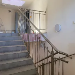 Montaż balustrady nierdzewnej na schodach w trakcie wykańczania wnętrza, widoczne niedokończone ściany, sufit z otworem i schody z płytkami.