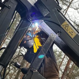 TECH-WELD Wojciech Putrym - Pracownik w masce spawalniczej i żółtych rękawicach spawa metalową konstrukcję na tle drzew, widoczne iskry i jasne światło spawania.