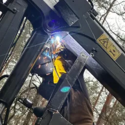 Pracownik w masce spawalniczej i żółtych rękawicach spawa metalową konstrukcję na tle drzew, widoczne iskry i jasne światło spawania.