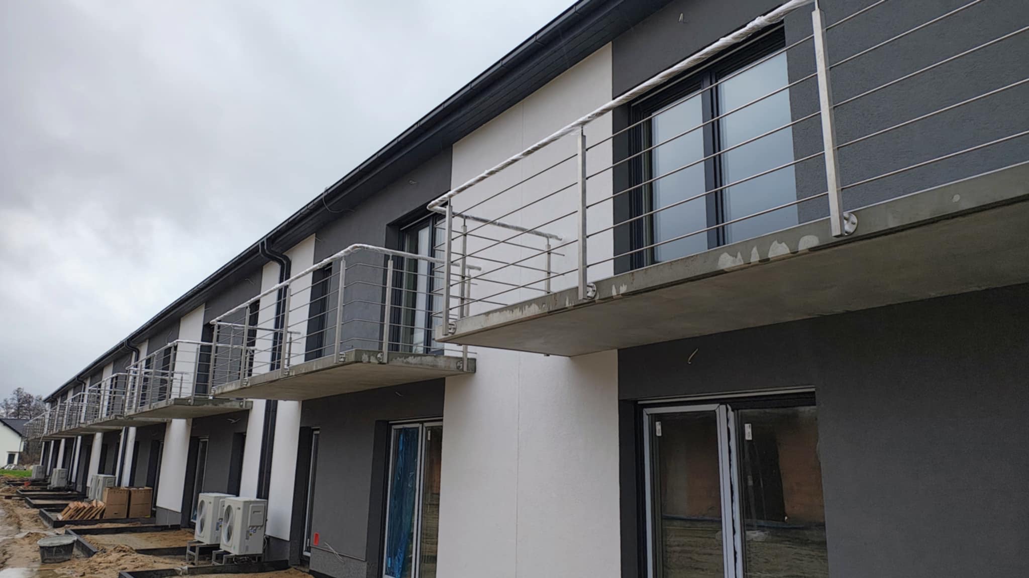 Rząd nowych budynków z balkonami zabezpieczonymi metalową konstrukcją, widoczne jednostki klimatyzacyjne na zewnątrz, teren budowy w trakcie wykończenia.