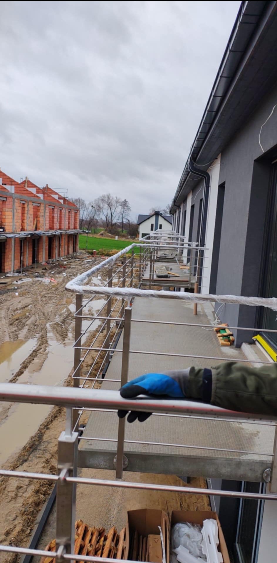 Montaż balustrady nierdzewnej na balkonie w trakcie budowy, widoczne błoto i niedokończone budynki w tle, pracownik w niebieskich rękawicach trzyma poręcz.