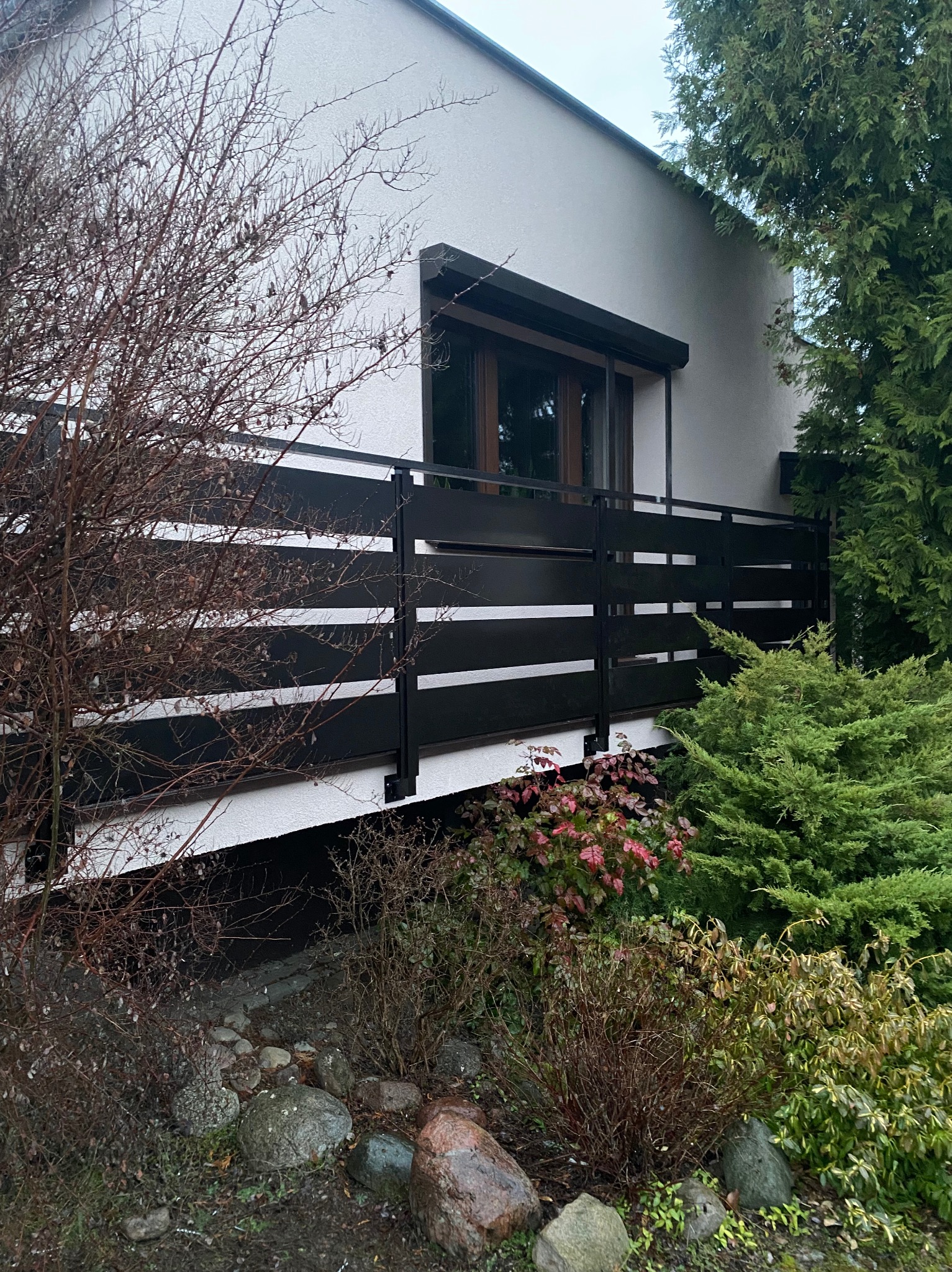 Czarna, pozioma balustrada balkonowa z metalowymi słupkami, widoczna z poziomu ogrodu, z krzewami i drzewami w tle.