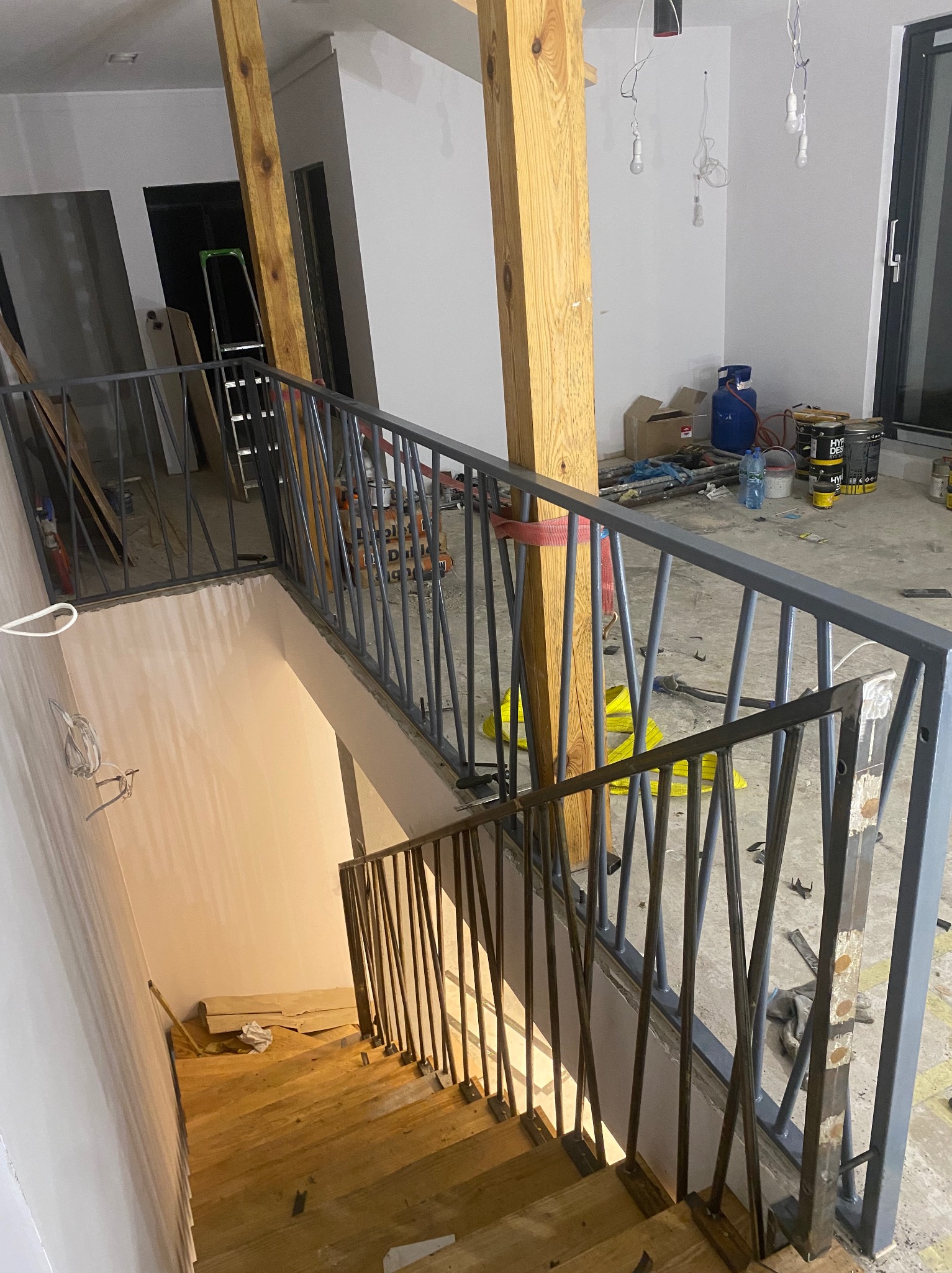 Wnętrze w trakcie wykańczania: drewniane schody z szarą metalową balustradą o pionowych prętach, widok z góry na dół, surowy wystrój, widoczne materiały budowlane i narzędzia.