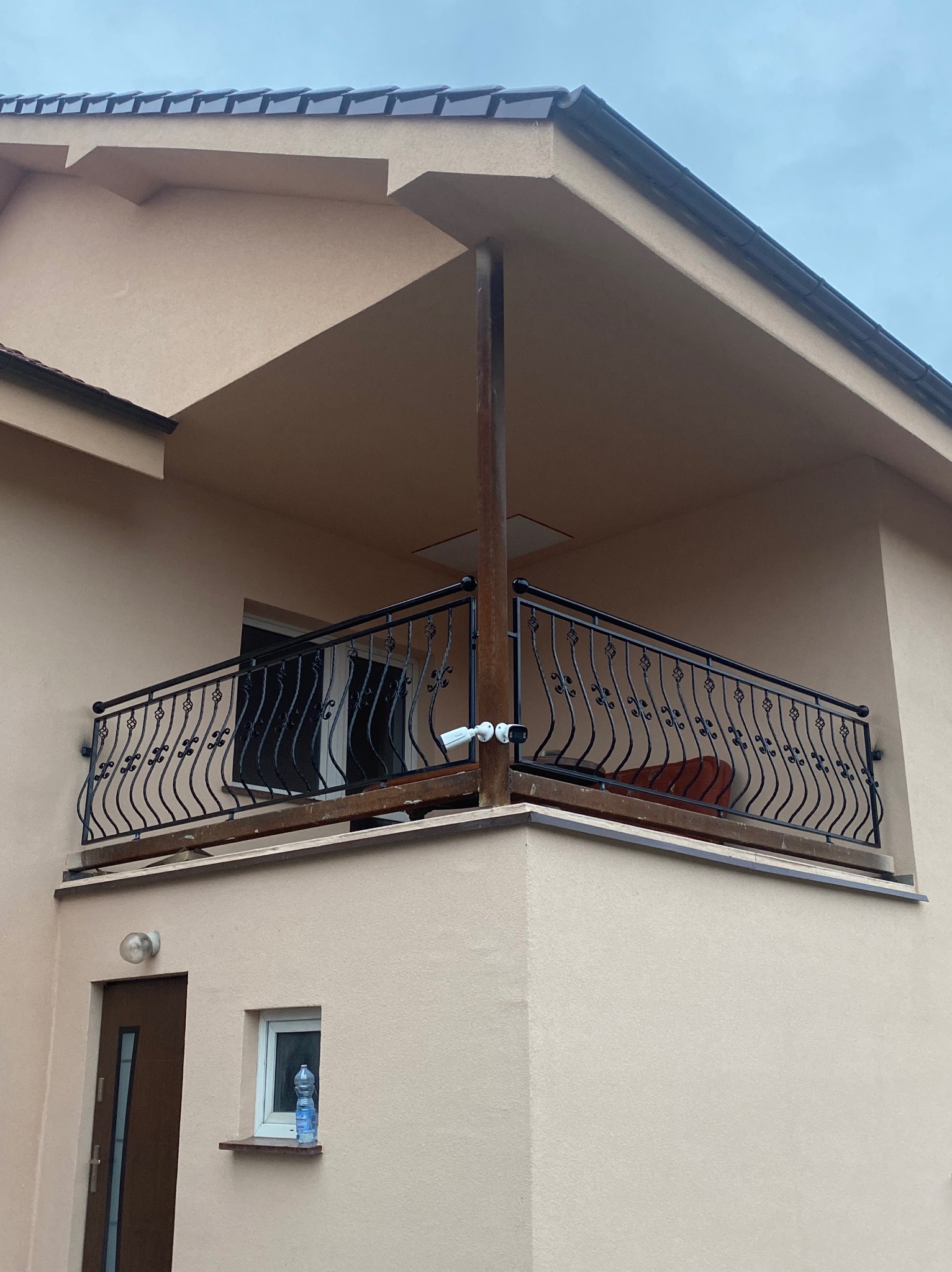 Czarna, metalowa barierka balkonowa z ozdobnymi elementami, zamontowana na balkonie domu o beżowej elewacji. Widoczna kamera monitoringu przymocowana do barierki.