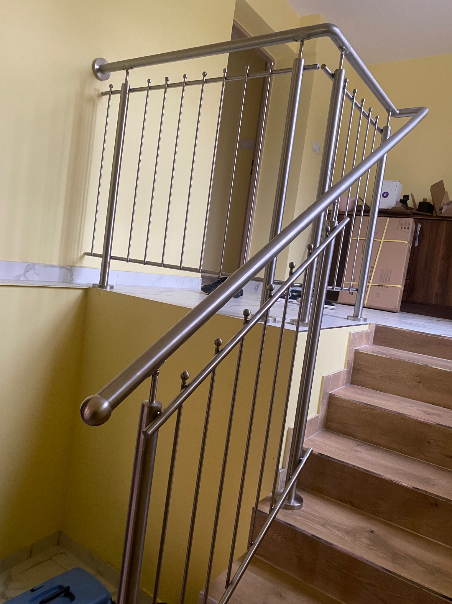 Wewnętrzna klatka schodowa z drewnianymi stopniami i nowoczesną, metalową barierką z pionowymi prętami, zakończonymi kulkami, na tle żółtej ściany.