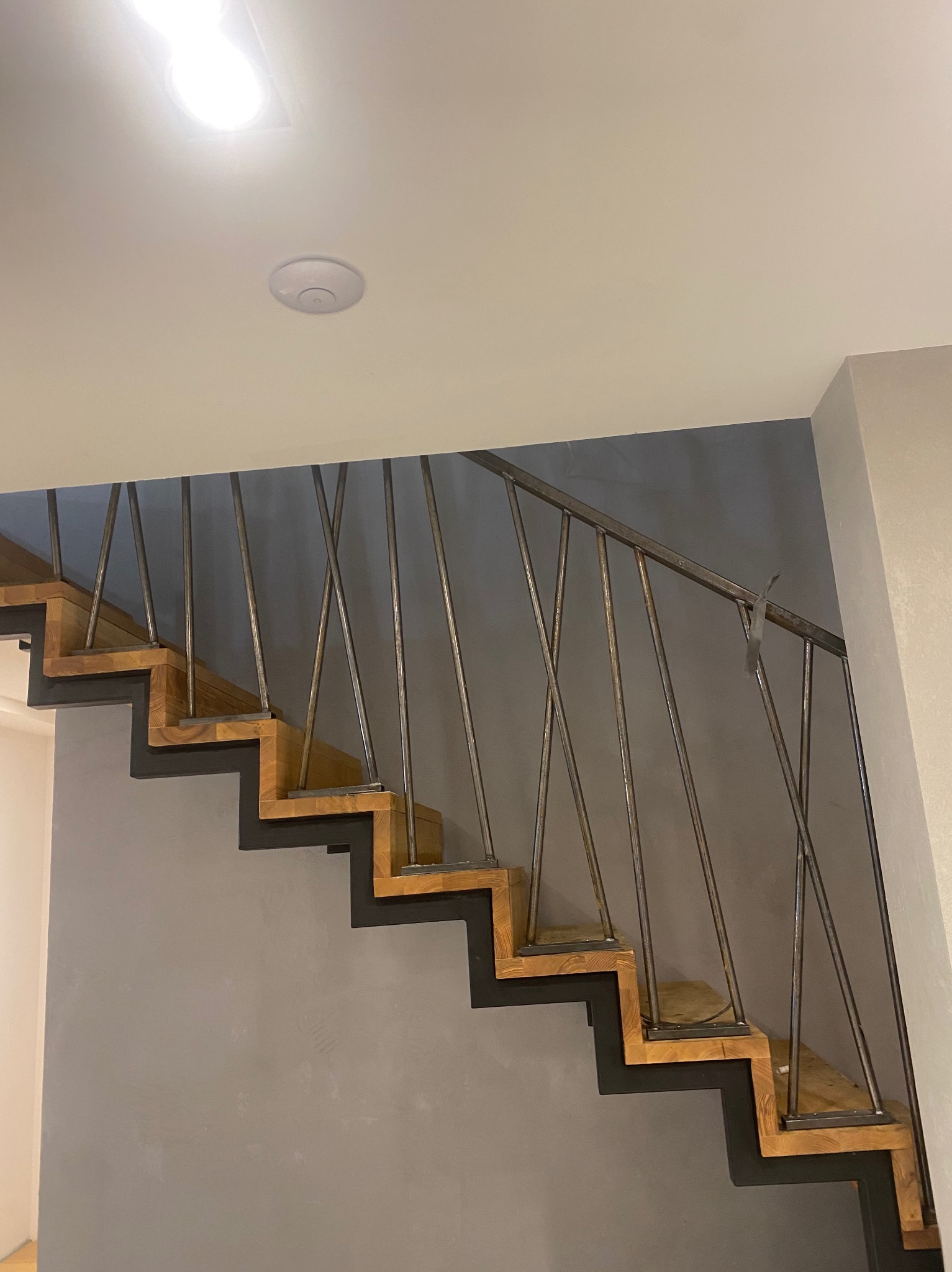 Nowoczesne schody z drewnianymi stopniami i metalową balustradą o minimalistycznym designie, widok z dołu na szarej ścianie, z taśmą przyklejoną do poręczy.