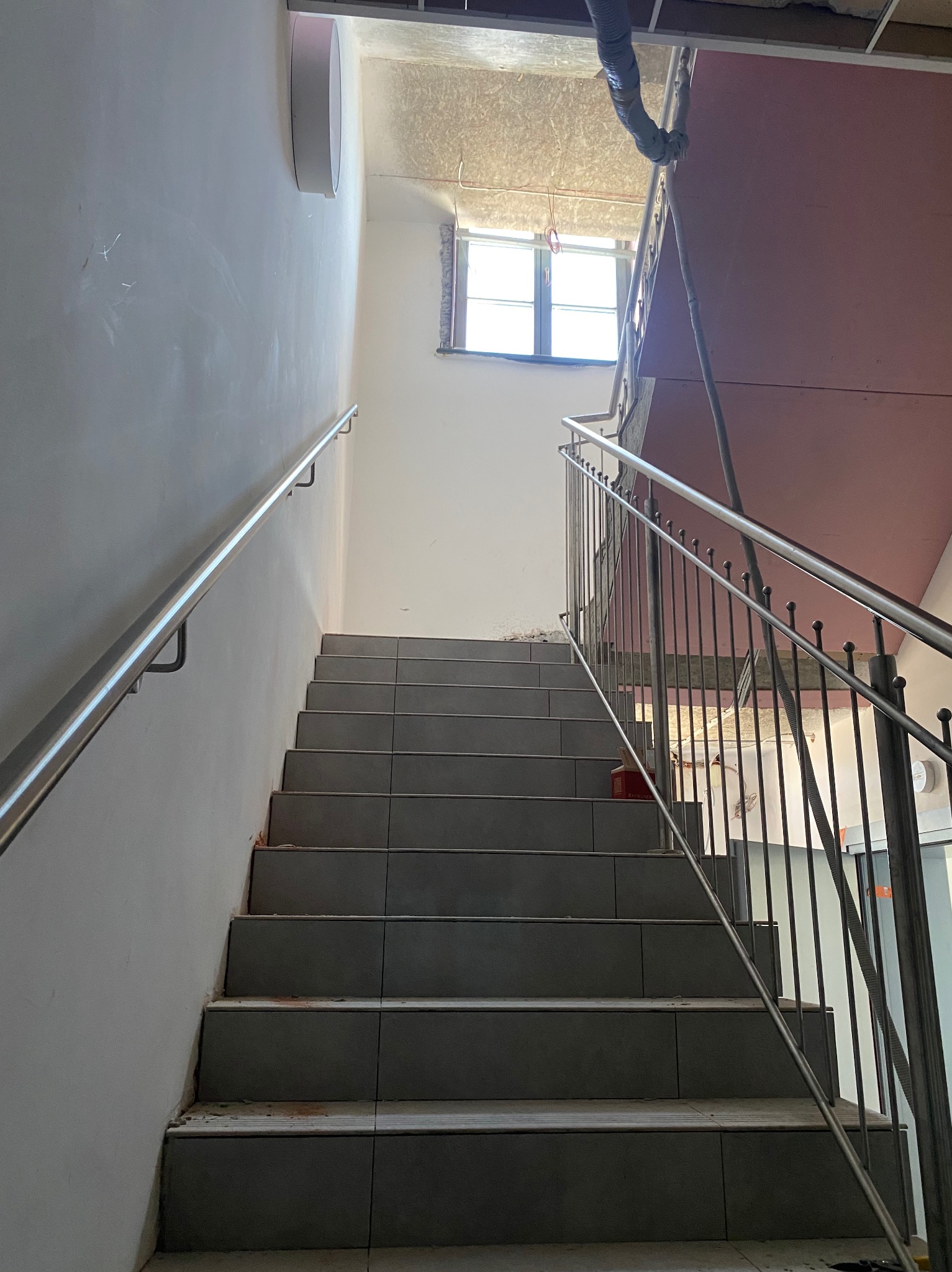 Wnętrze klatki schodowej w trakcie wykańczania, widoczne szare płytki na schodach, metalowa barierka z pionowymi prętami i okrągłymi zakończeniami, oraz rura wentylacyjna pod sufitem.
