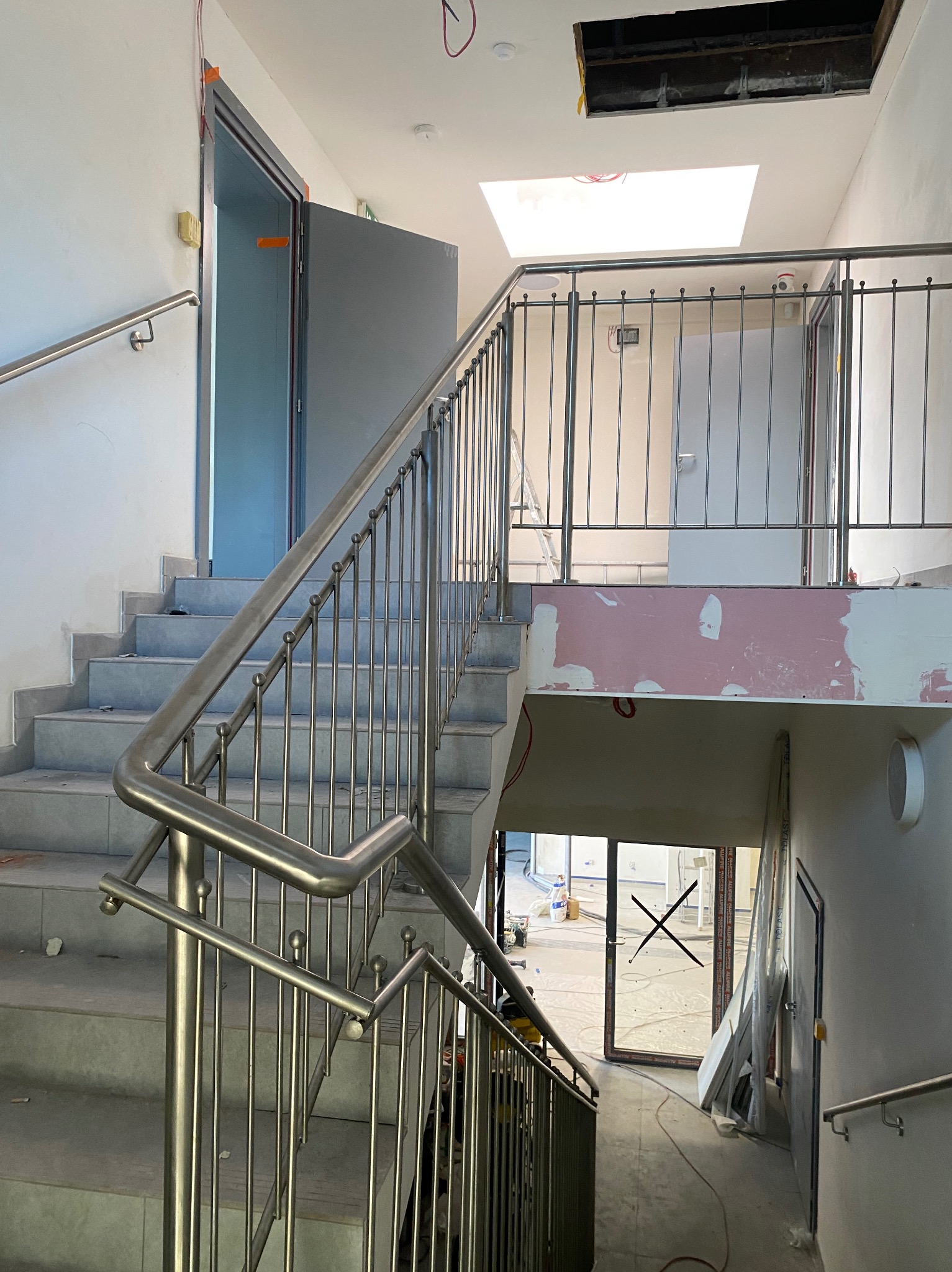 Klatka schodowa w trakcie wykończenia, widoczne schody wyłożone szarymi płytkami, metalowe barierki z pionowymi prętami i okrągłymi zakończeniami, surowy stan ścian, otwór w suficie i okno dachowe.