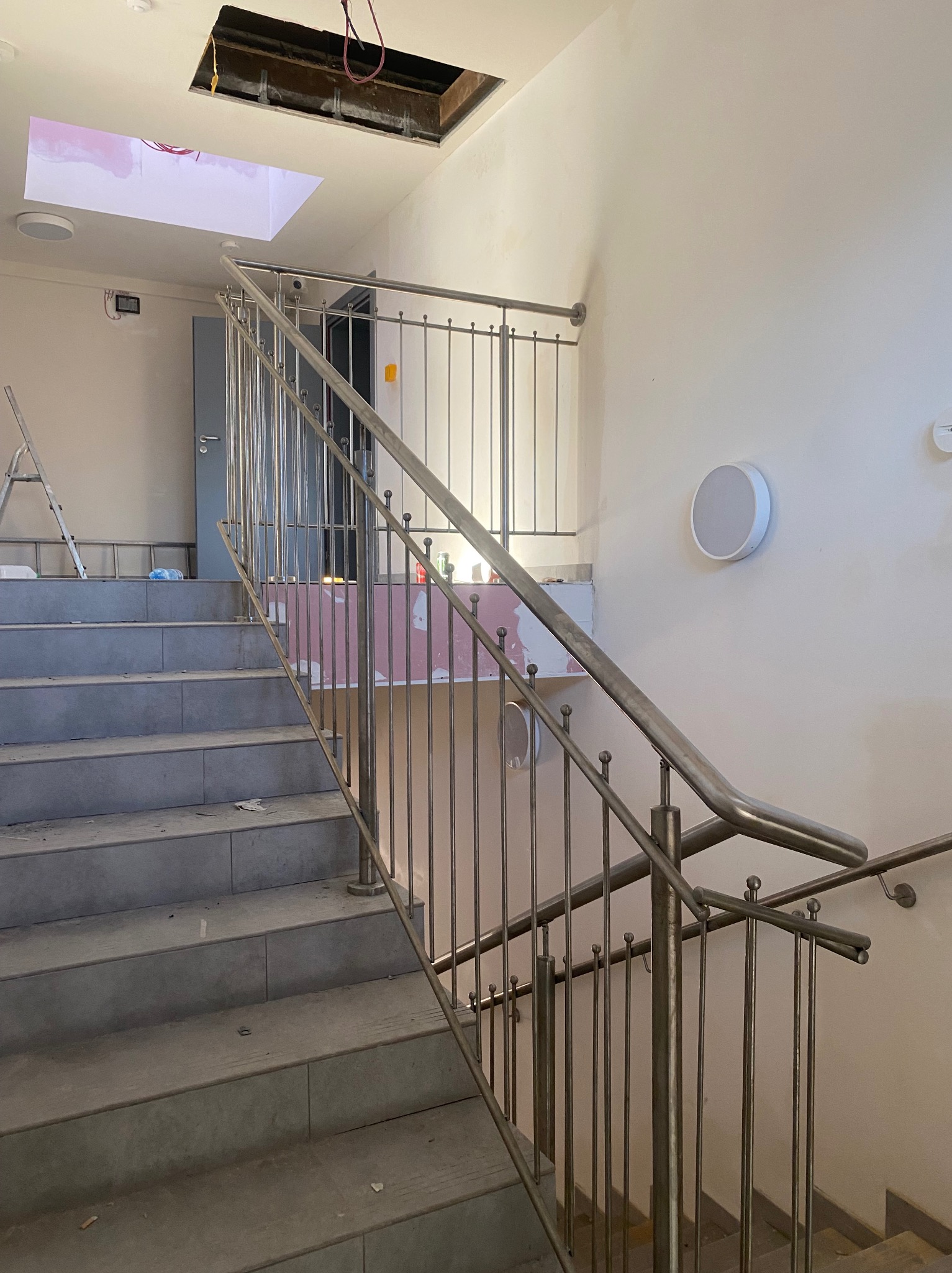 Montaż balustrady nierdzewnej na schodach w trakcie wykańczania wnętrza, widoczne niedokończone ściany, sufit z otworem i schody z płytkami.