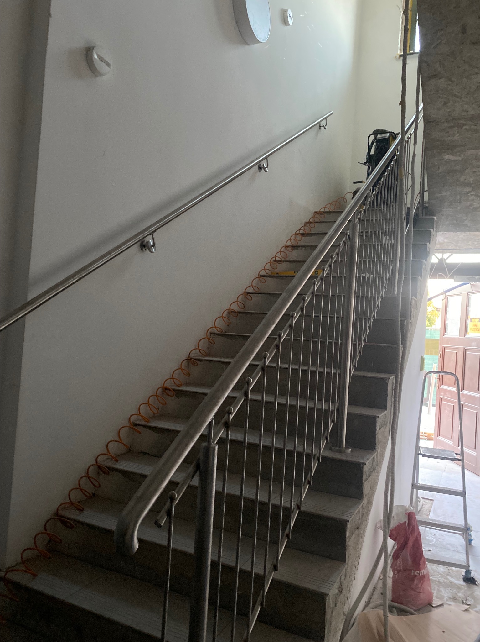 Montaż barierki ze stali na schodach betonowych wewnątrz budynku, widoczny zwój pomarańczowego przewodu, drabina i worki z zaprawą.