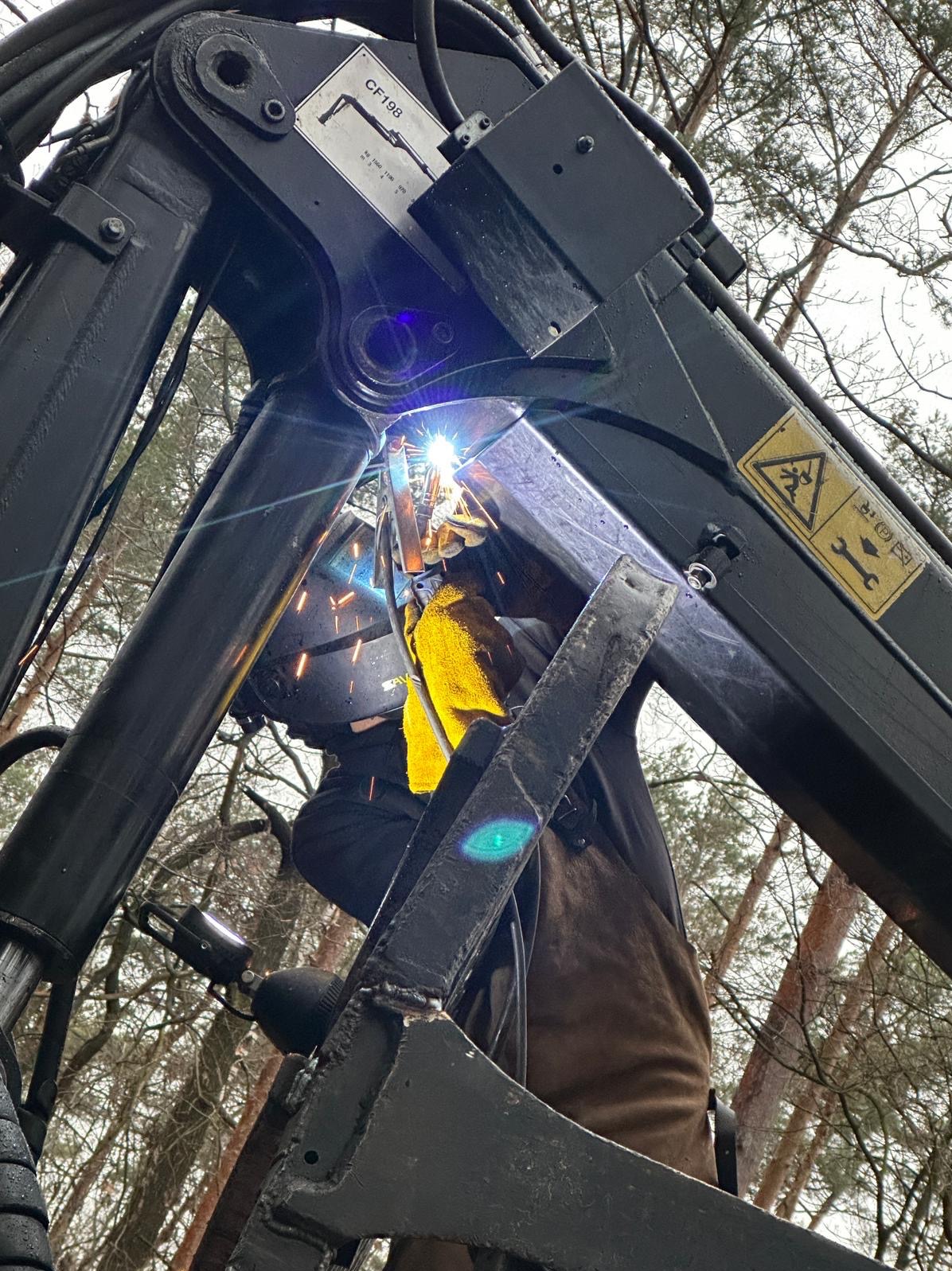 Pracownik w masce spawalniczej i żółtych rękawicach spawa metalową konstrukcję na tle drzew, widoczne iskry i jasne światło spawania.