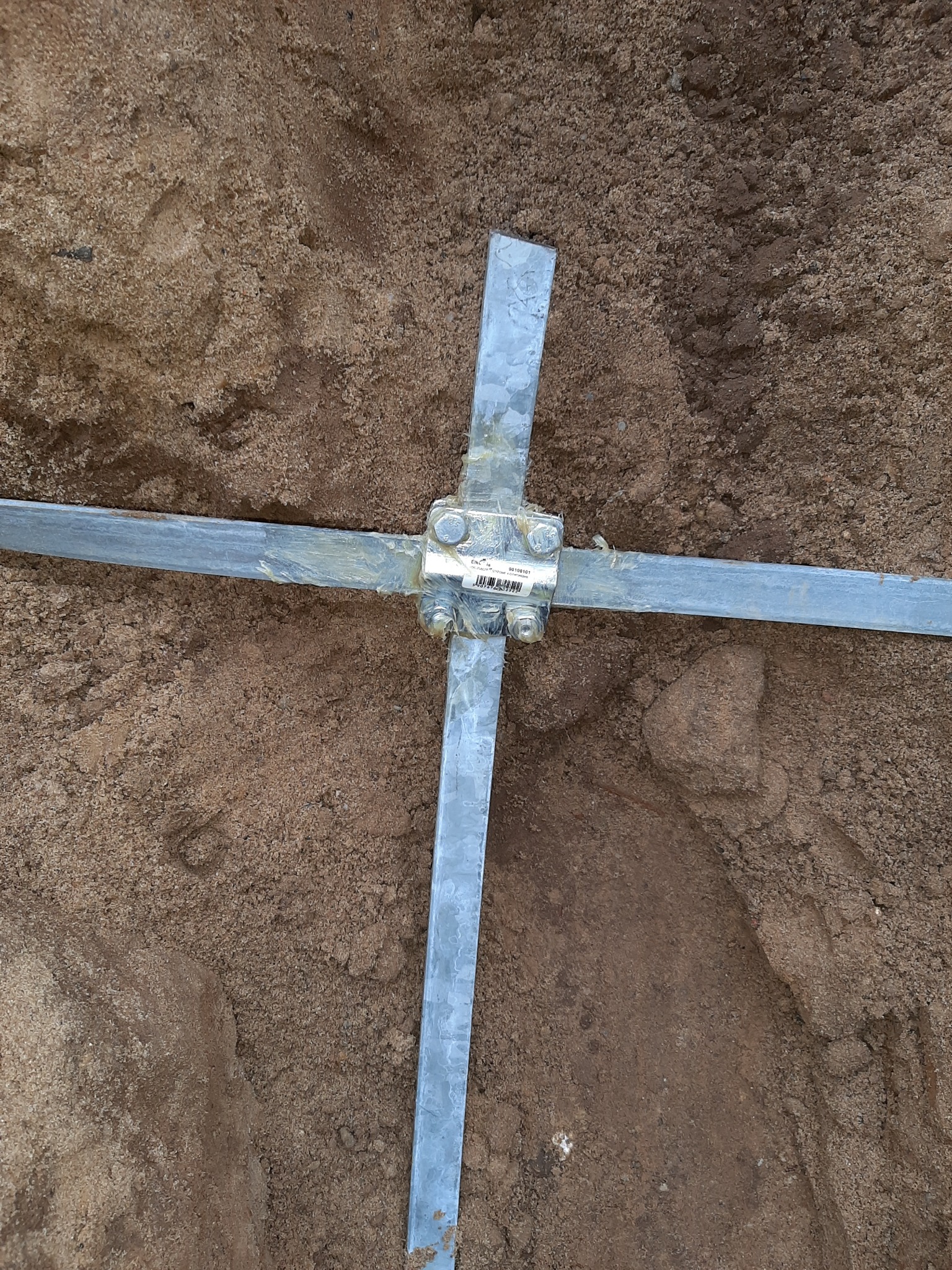 Połączenie krzyżowe ocynkowanych bednarek uziemiających w wykopie, widoczne ślady świeżego montażu i zabrudzenia ziemią.