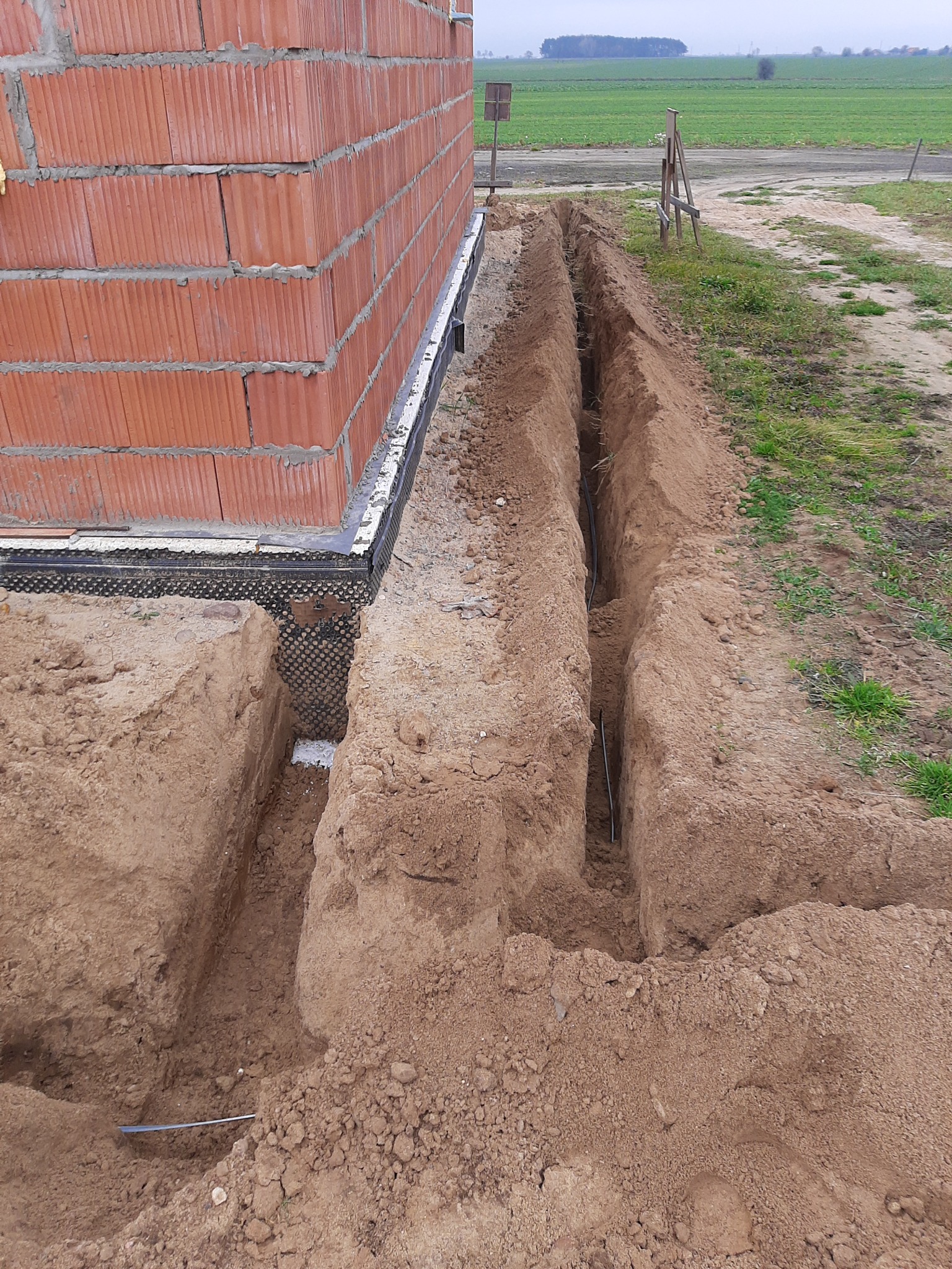 Wykopane wąskie rowy w ziemi wzdłuż fundamentów budynku z czerwonej cegły, widoczne rury instalacyjne w rowach, tło to pole i pochmurne niebo.