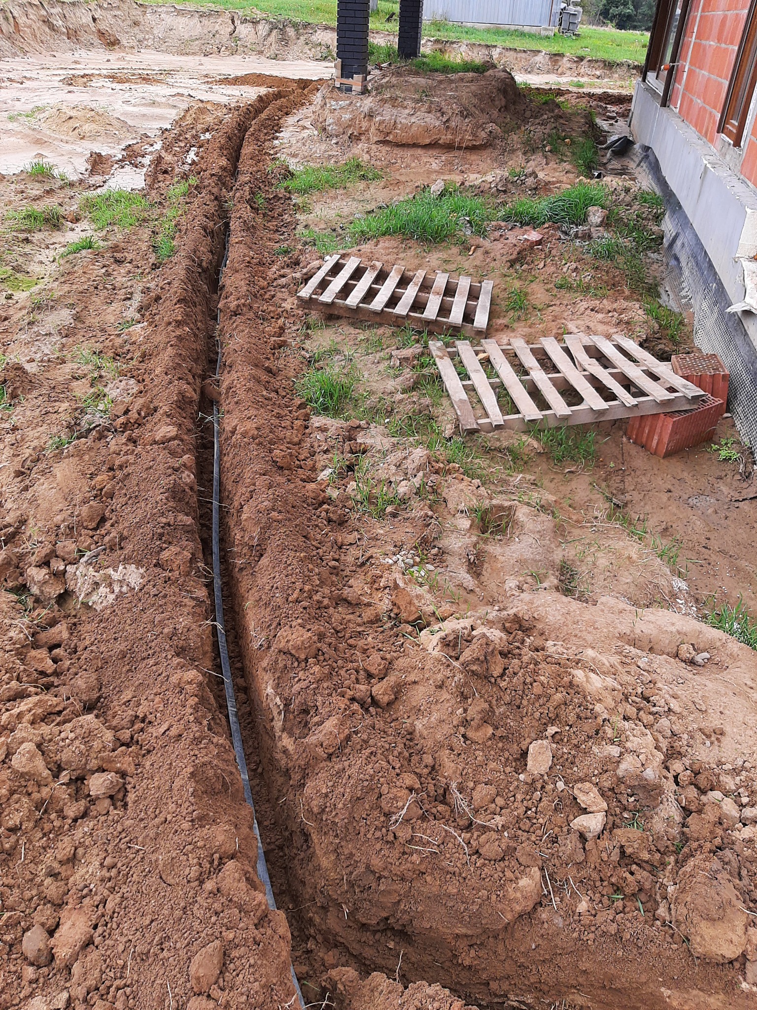 Wykopany wąski rów w ziemi z ułożoną czarną rurą, widoczne palety i fragment ściany budynku w trakcie budowy z cegły.