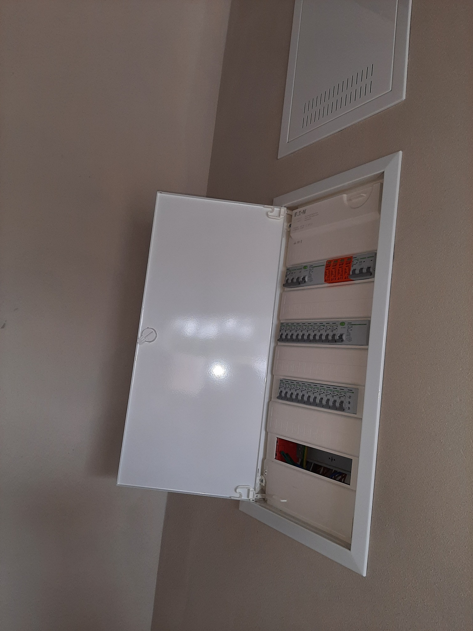 Otwarta biała skrzynka rozdzielcza Eaton na beżowej ścianie, widoczne bezpieczniki i okablowanie, obok kratka wentylacyjna.