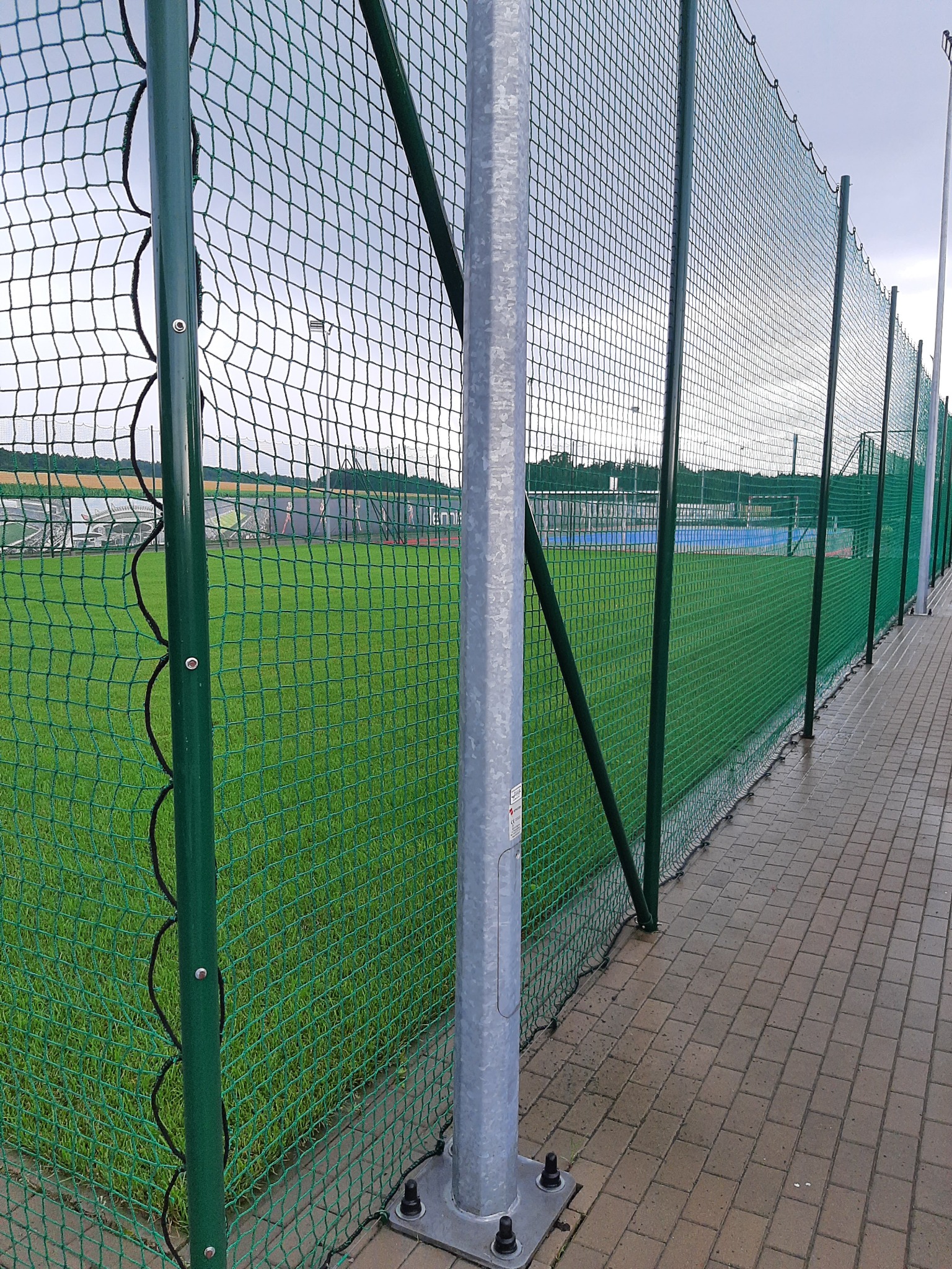 Ochronna siatka z zielonymi słupkami okalająca boisko sportowe ze sztuczną nawierzchnią, widoczny fragment chodnika z kostki brukowej.