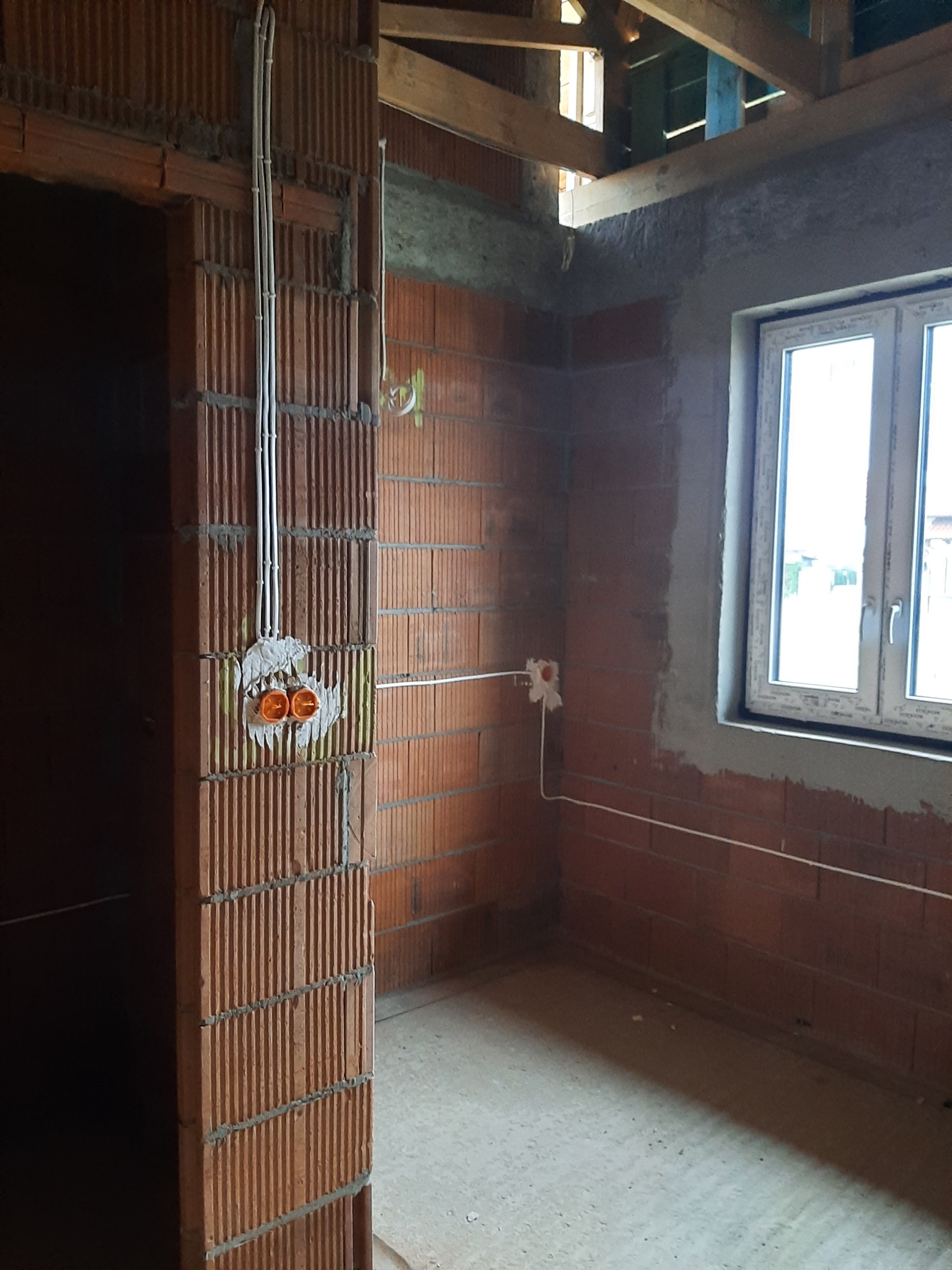 Wnętrze w trakcie prac wykończeniowych, widoczne przewody elektryczne prowadzone po ceglanej ścianie, puszki pod gniazdka w stanie surowym, okno z białymi ramami.