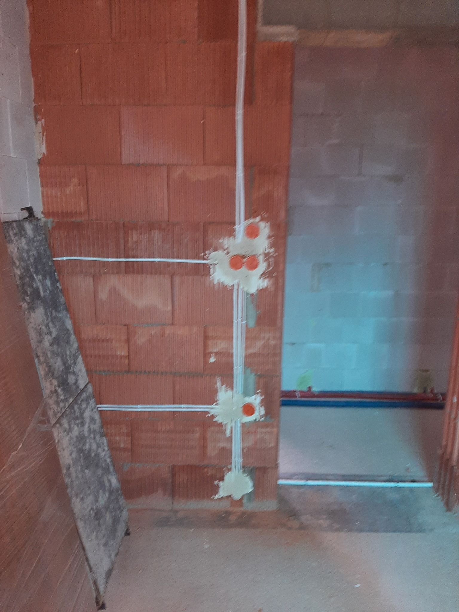 Wewnętrzna instalacja elektryczna w surowym budynku: białe przewody i puszki montażowe z pomarańczowymi elementami na ceglanej ścianie, widok na sąsiednie pomieszczenie.