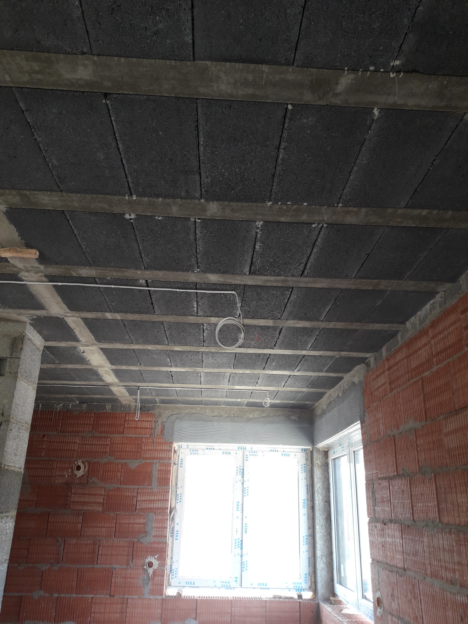 Wnętrze w trakcie budowy: widok na strop z betonowych pustaków i częściowo wymurowane ściany z czerwonej cegły dziurawki, instalacja elektryczna w peszlu zwisająca ze stropu.