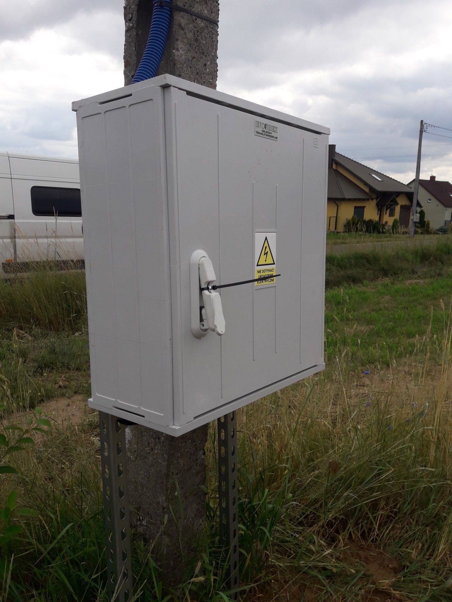 Szara skrzynka elektryczna zamontowana na betonowym słupie z metalowymi wspornikami, otoczona trawą i z ostrzeżeniem o wysokim napięciu.