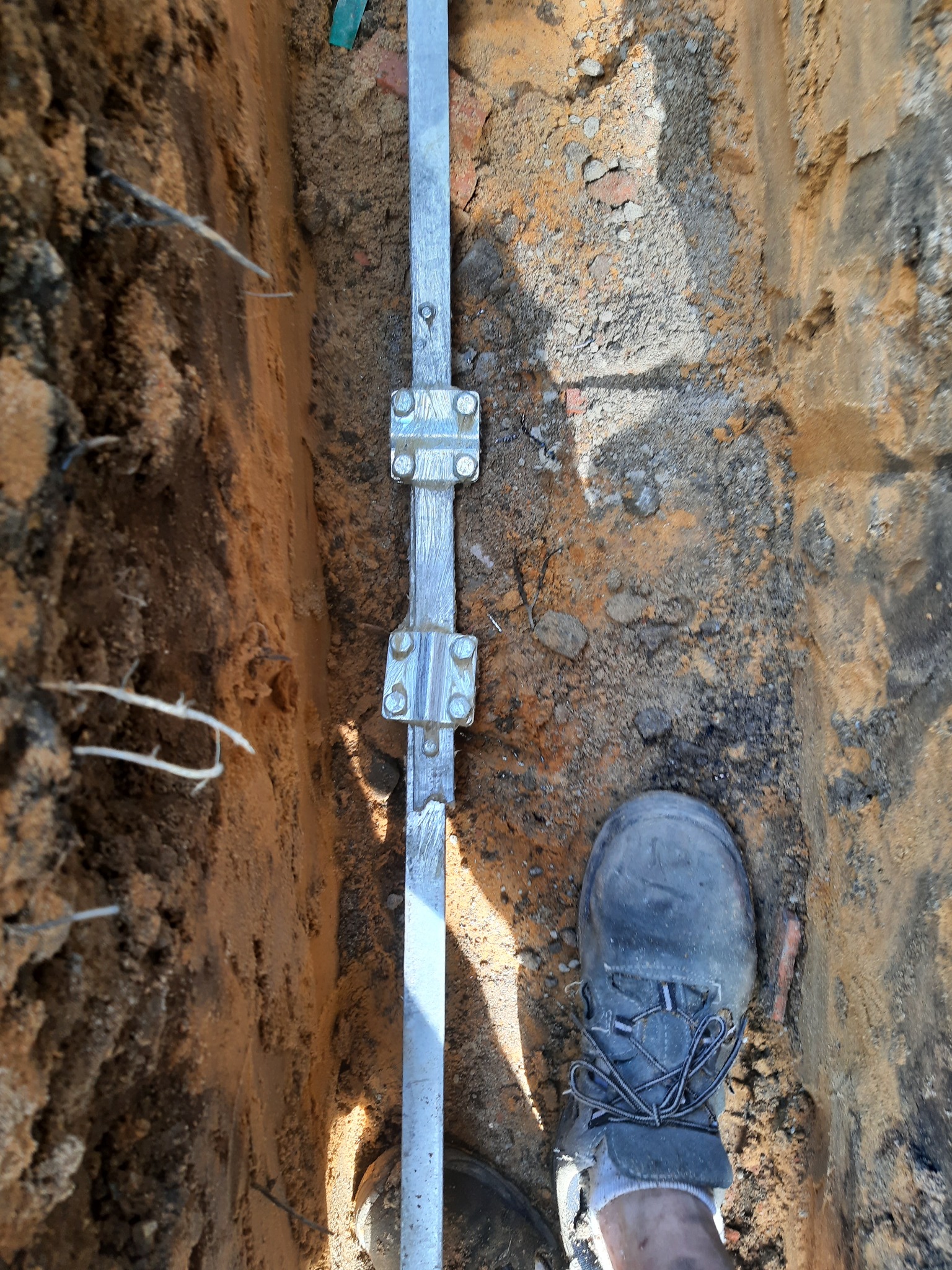 Metalowy uziom z łącznikami śrubowymi w wykopie ziemnym, widoczny fragment roboczego buta dla skali.