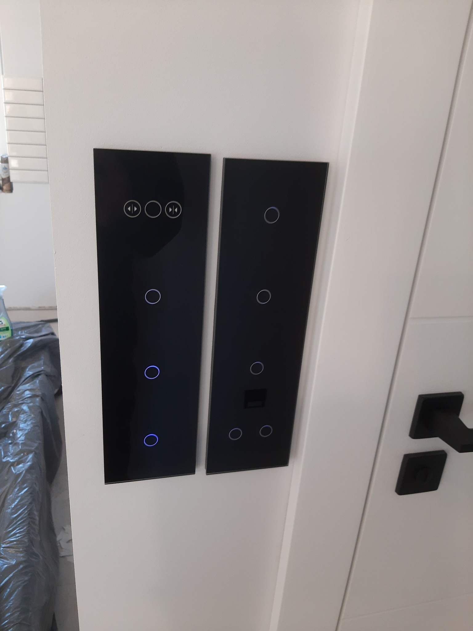 Dwa czarne, dotykowe panele sterowania oświetleniem z niebieskimi podświetleniami, zamontowane na białej ścianie obok białych drzwi z czarną klamką.