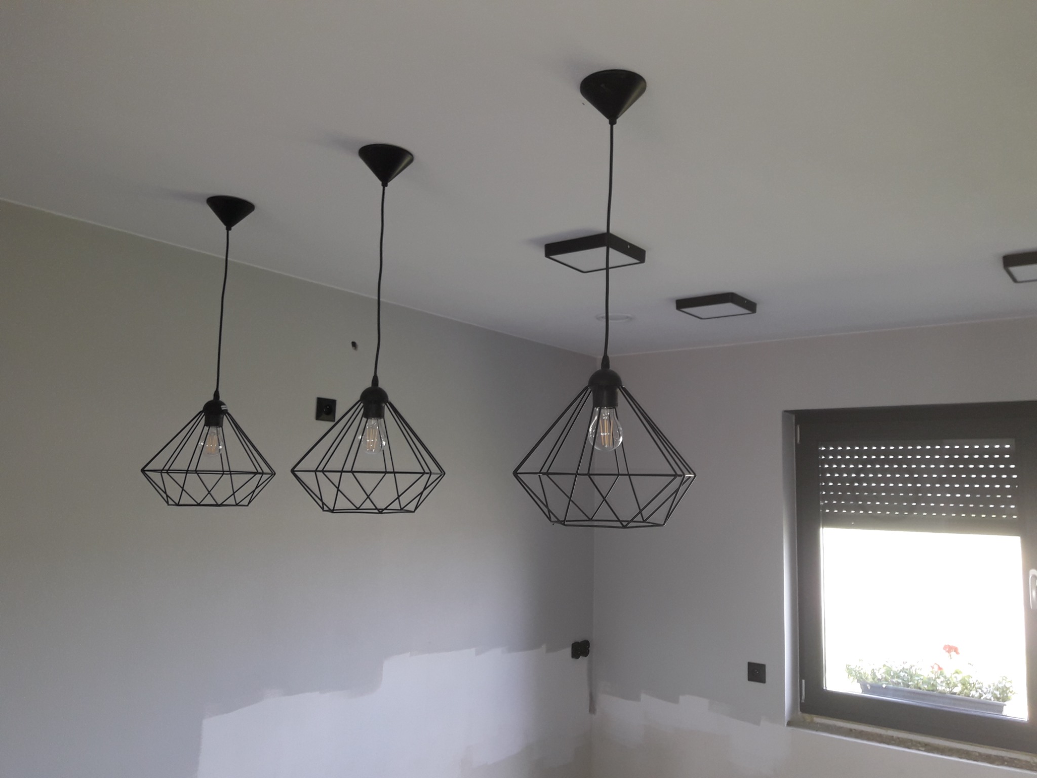 Trzy czarne, geometryczne lampy wiszące z żarówkami Edisona na tle szarej ściany i okna z żaluzjami.
