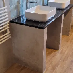 Nowoczesna łazienka z dwiema umywalkami nablatowymi na czarnym blacie, podwieszana toaleta i grzejnik drabinkowy na tle jasnych, pionowo ułożonych płytek.