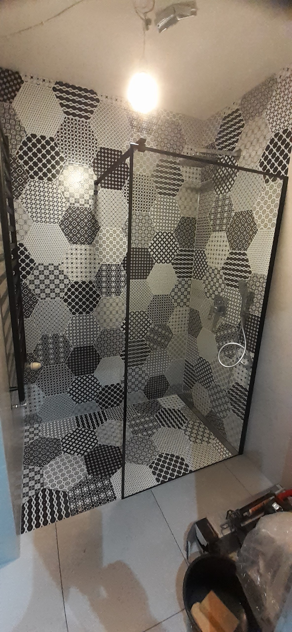 Ukończona łazienka z kabiną prysznicową, wyłożona heksagonalnymi płytkami w czarno-białe wzory, widoczne narzędzia i wiadro w rogu.