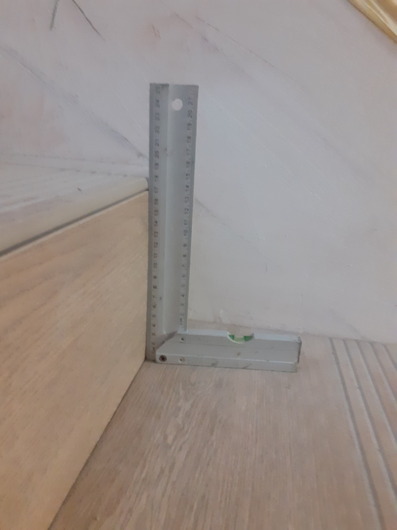 Kątownik stolarski z poziomicą przyłożony do krawędzi panela podłogowego i ściany, sprawdzanie kąta prostego podczas montażu.