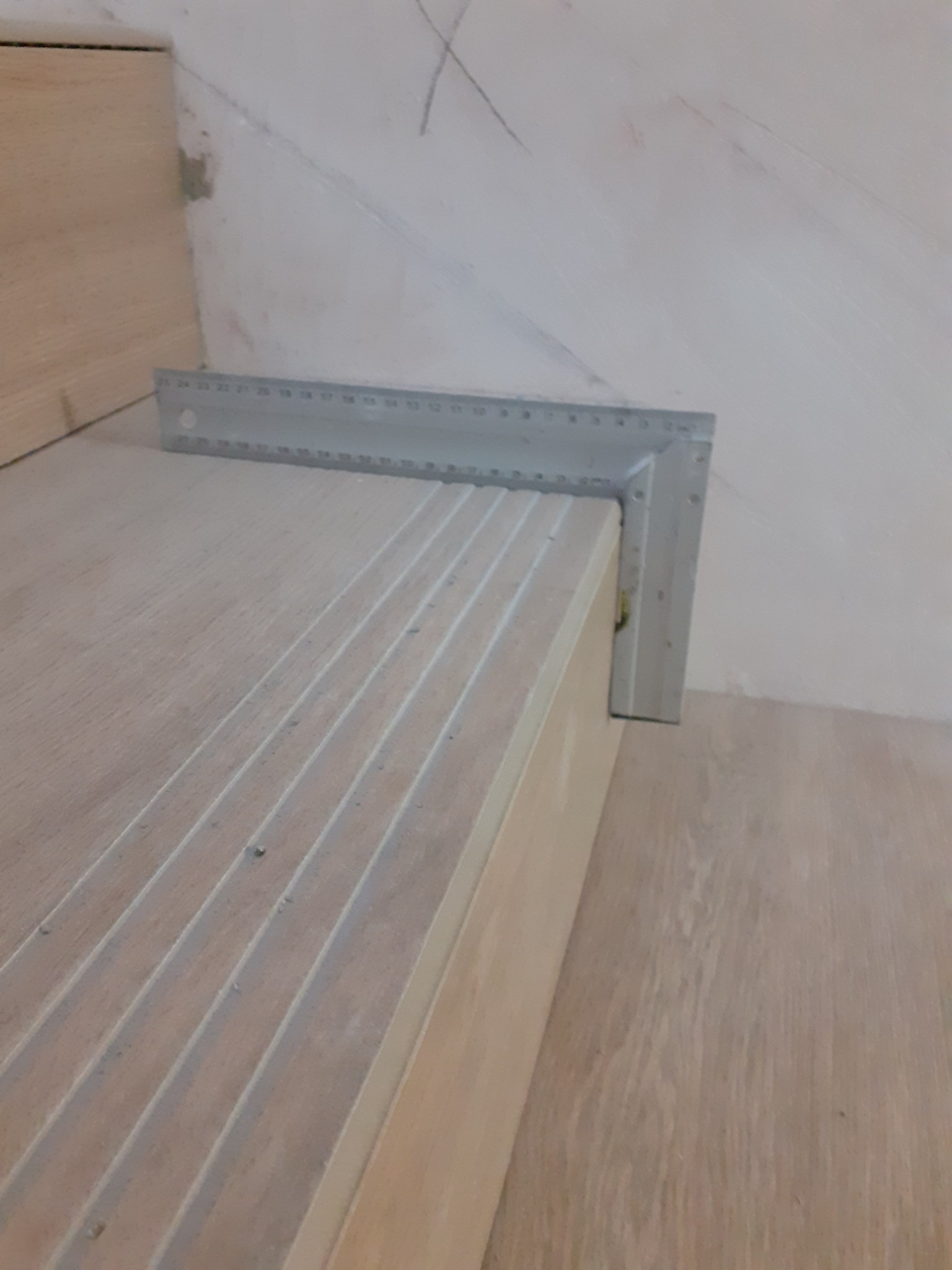 Ułożony drewniany stopień schodów z rowkami, sprawdzany kątownikiem na tle surowej ściany z widocznymi śladami gładzi.