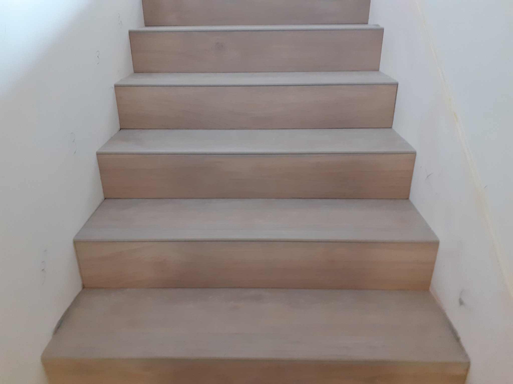 Schody wykończone jasnym drewnem, widok z dołu, ściana z drobnymi zabrudzeniami w tle.