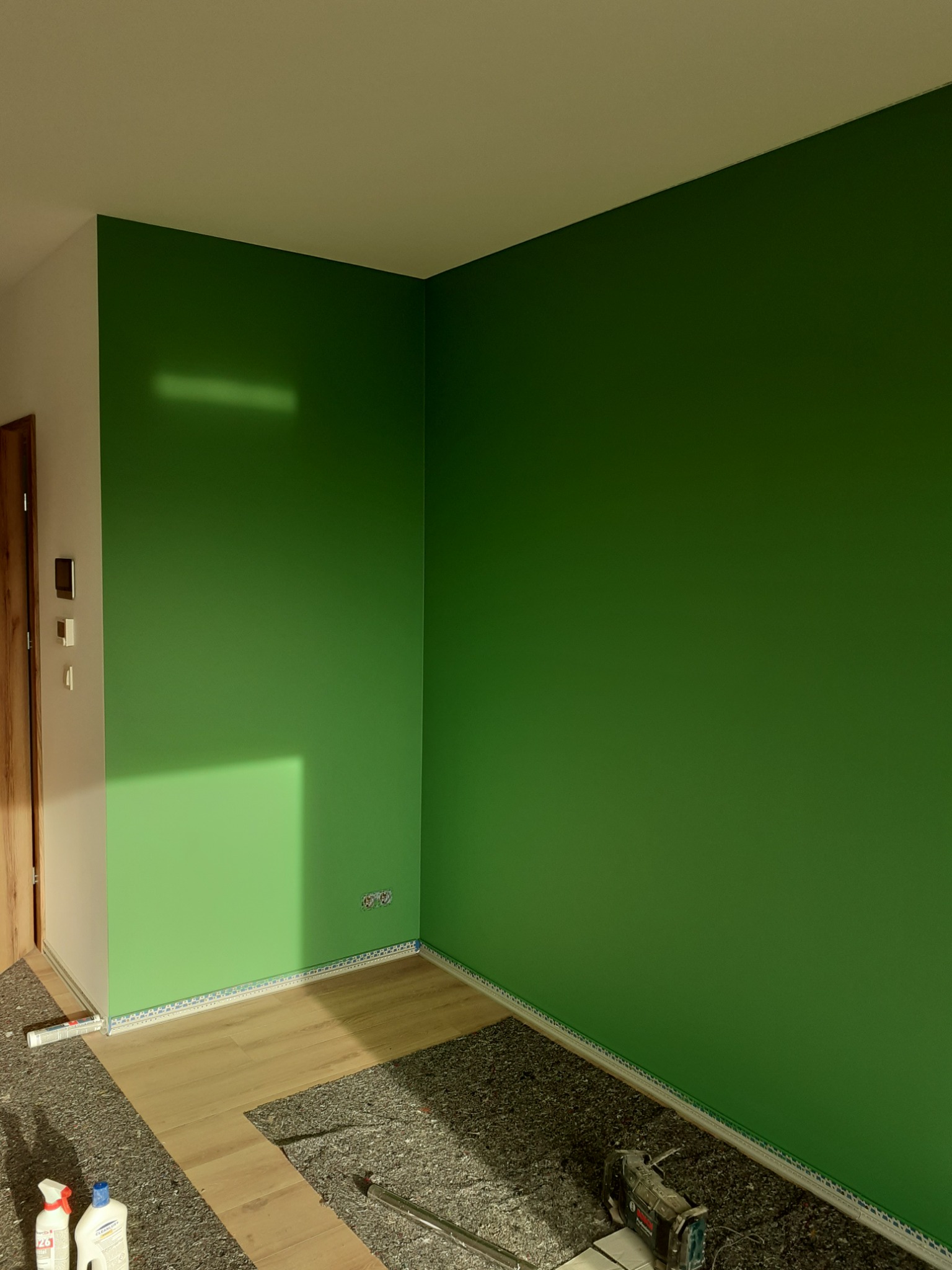 Pokój z zielonymi ścianami w trakcie układania parkietu, widoczna warstwa izolacyjna, listwy przypodłogowe i narzędzia.