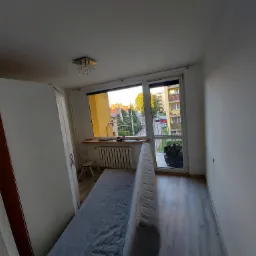 Pusty pokój z białymi ścianami i parkietem, widok na balkon i osiedle za oknem, widoczna szafa z brązowymi wstawkami, mały taboret i grzejnik pod oknem, sufit z lampą kryształową.