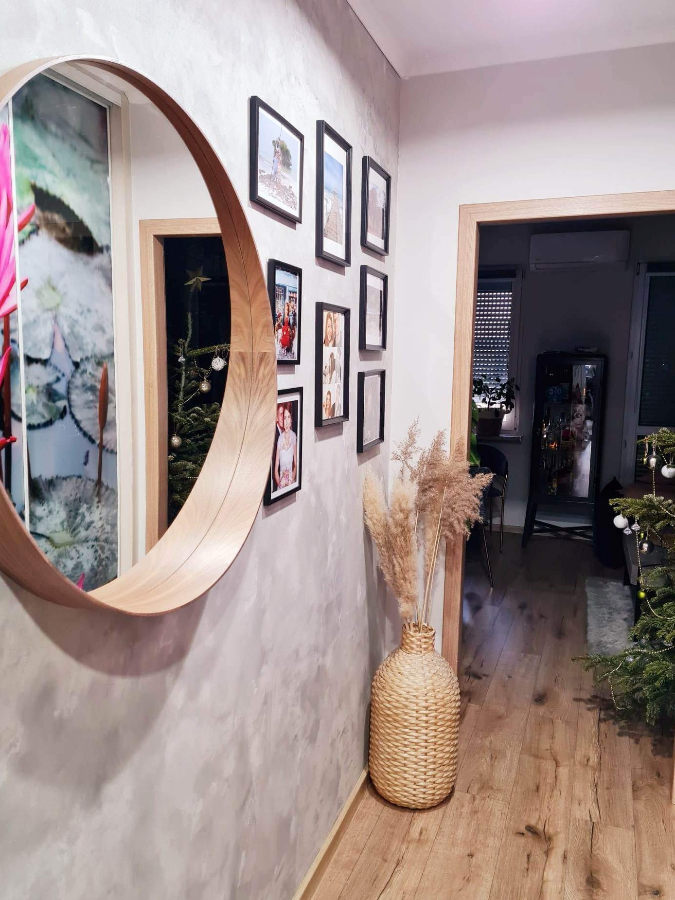 Hol wejściowy po odświeżeniu: ściana z efektem betonu, okrągłe lustro z drewnianą ramą, galeria zdjęć, dekoracyjny wazon z suszem, drewniana podłoga i choinka w tle.