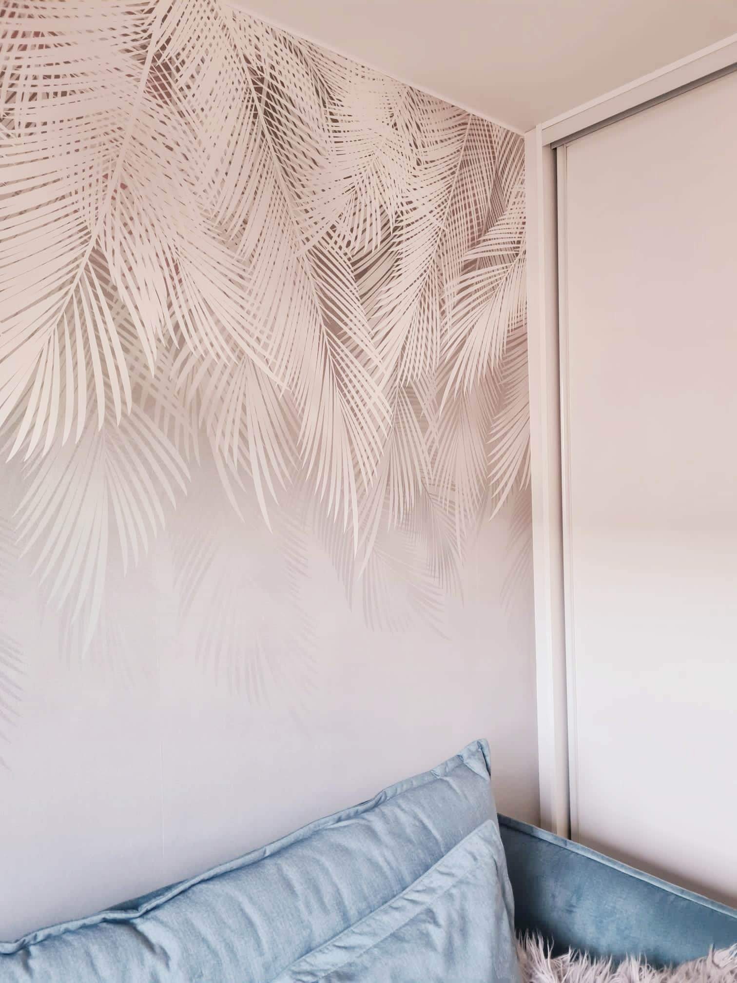 Ściana z tapetą w motywie jasnych liści palmowych, przylegająca do białej szafy przesuwnej, z fragmentem niebieskiego zagłówka łóżka w dolnej części kadru.