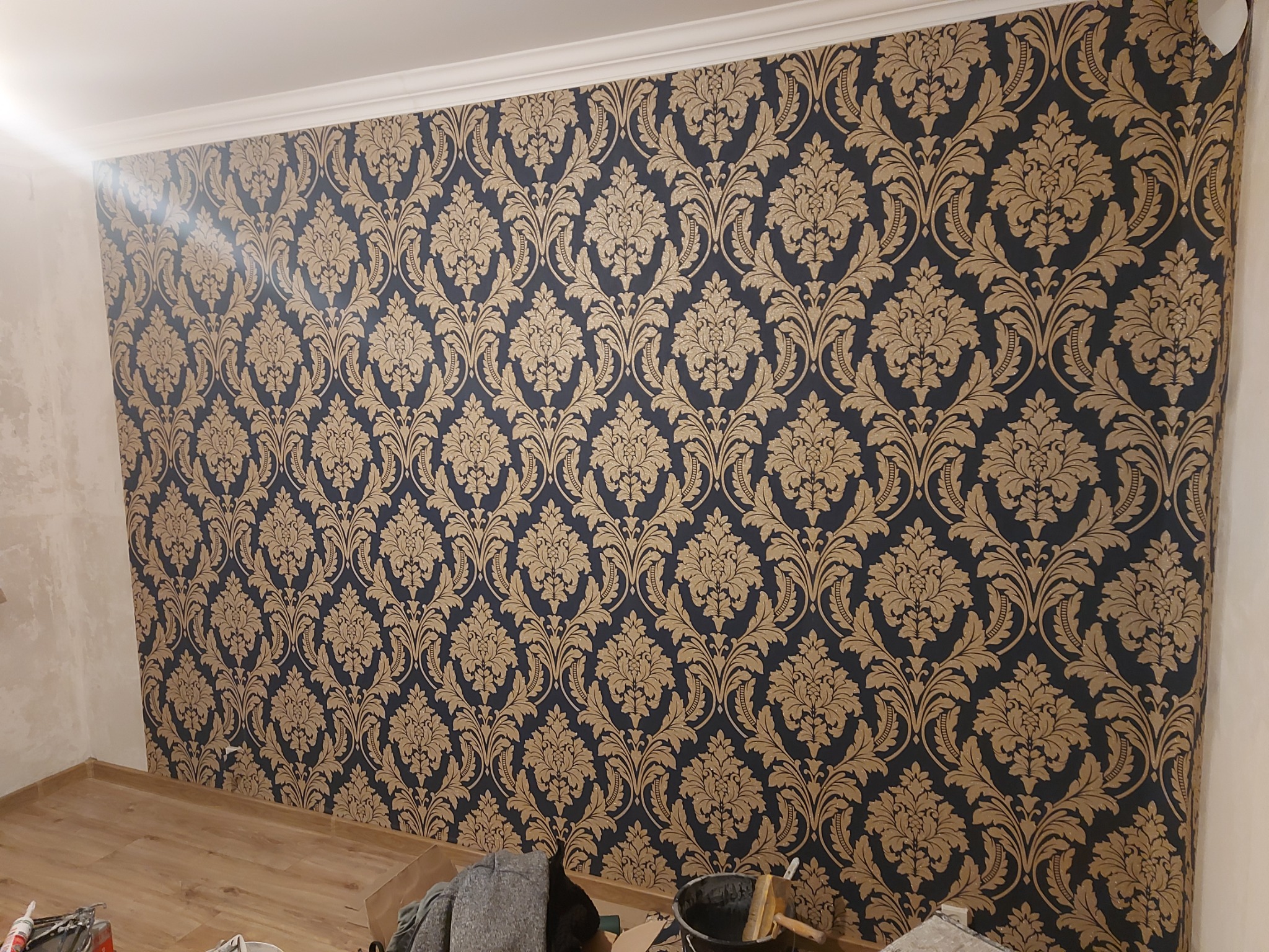 Ściana pokryta tapetą w ciemnogranatowe tło z powtarzającym się złotym motywem roślinnym; widoczne narzędzia malarskie i wiadro z farbą przy podłodze.
