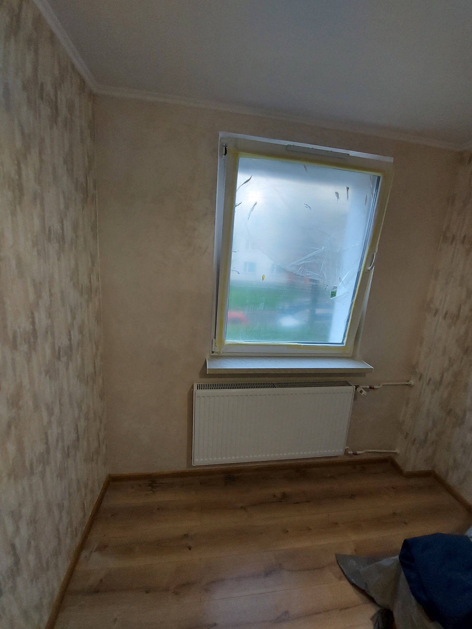 Pokój w trakcie prac przygotowawczych do malowania: okno zabezpieczone folią malarską, ściany z efektem dekoracyjnym, grzejnik zamontowany, podłoga drewniana.