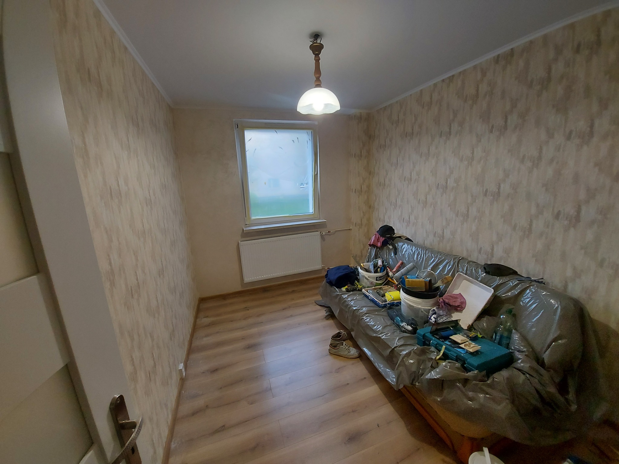 Pokój w trakcie remontu: ściany z tapetą w odcieniach beżu, podłoga z paneli, okno z zasłoniętą szybą, sofa przykryta folią z narzędziami malarskimi i wiaderkami z farbą, wisząca lampa z białym...