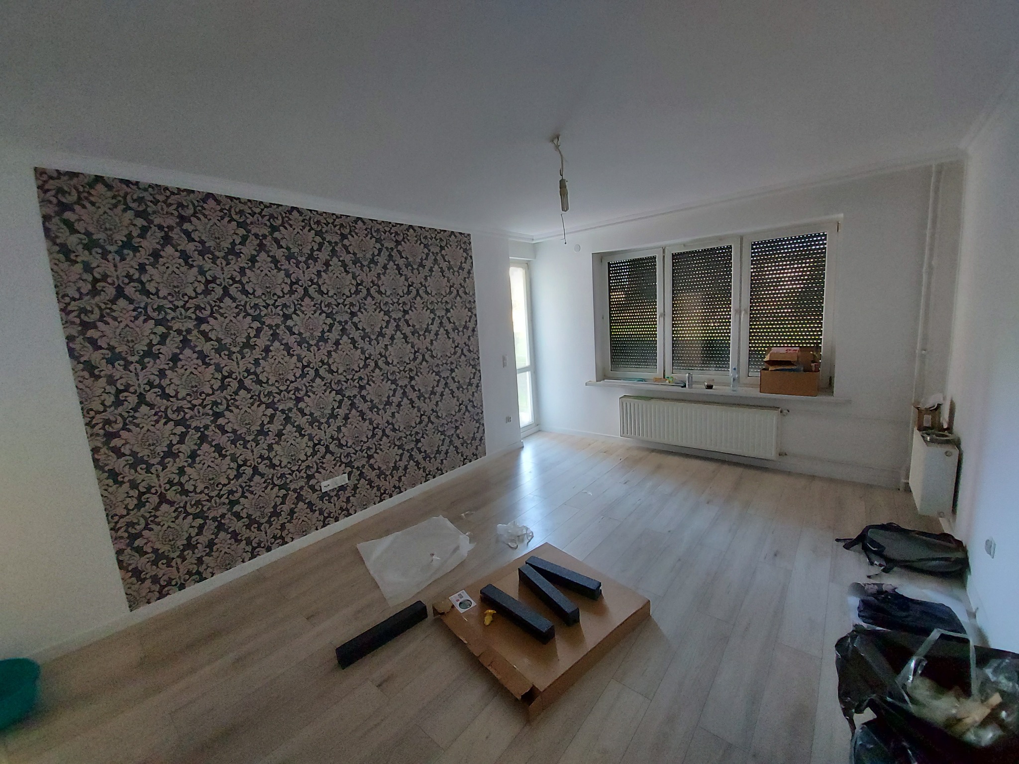 Pokój w trakcie prac: jedna ściana z ciemną tapetą w kwiaty, reszta ścian biała, drewnopodobna podłoga, okno z żaluzjami, karton z elementami montażowymi na podłodze, worki na śmieci przy ścianie.