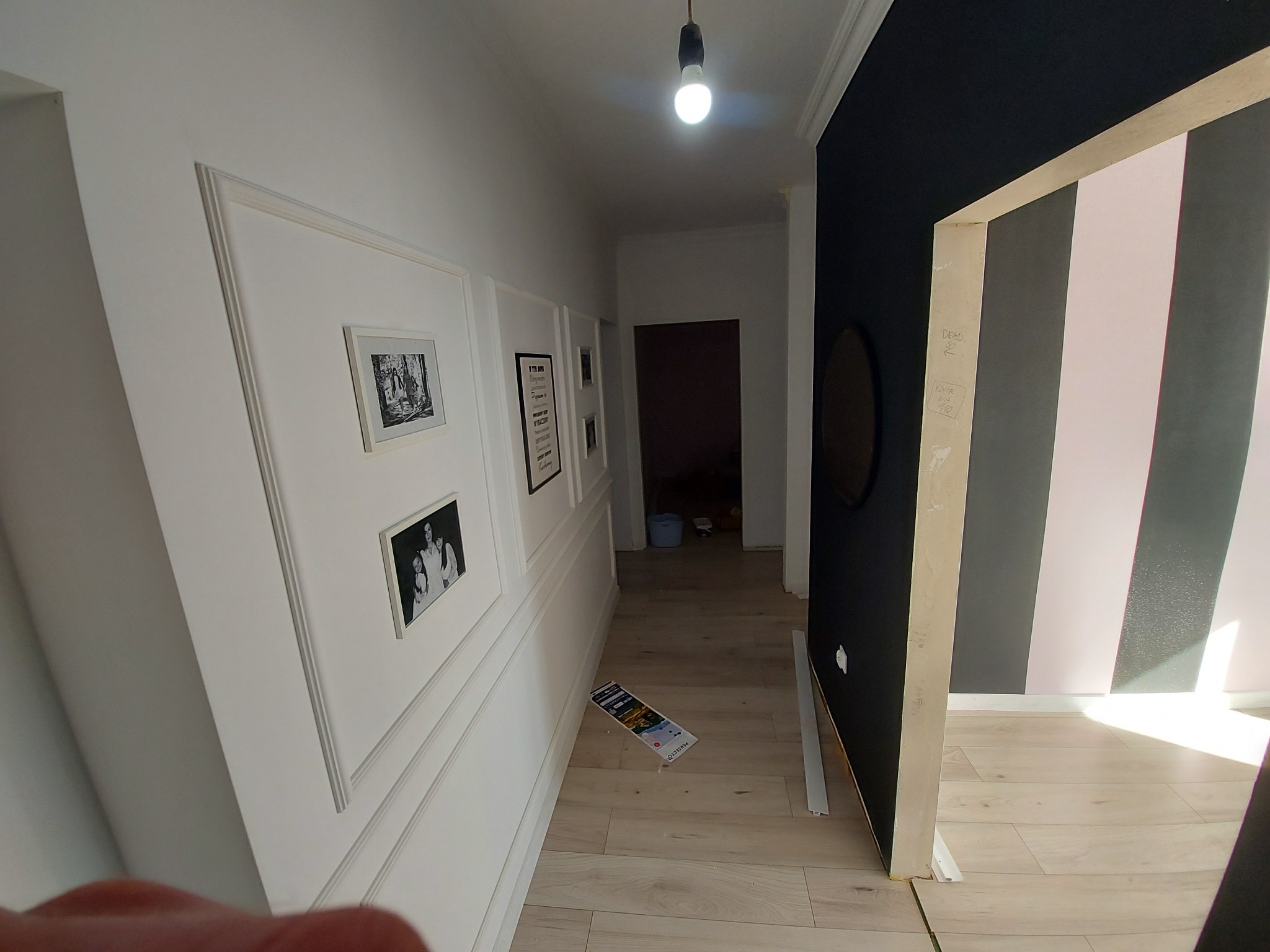 Korytarz po remoncie z panelami ściennymi, ramkami na zdjęcia, pomalowaną ścianą w pasy i drewnianą podłogą, widoczny prospekt na podłodze.