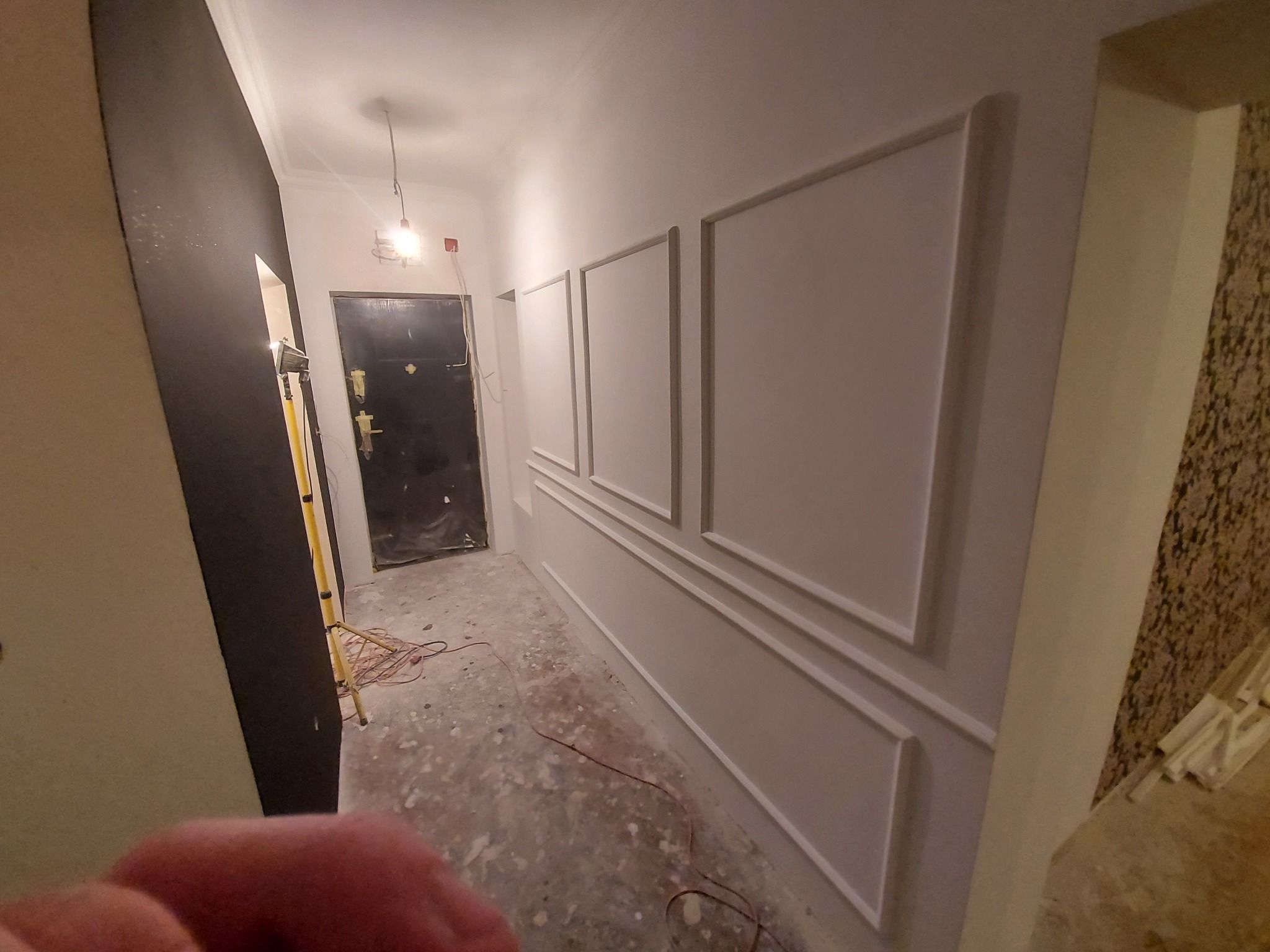 Korytarz w trakcie prac wykończeniowych: ściany pomalowane na biało z dekoracyjnymi listwami, surowa podłoga, widoczne przewody elektryczne i lampa robocza.
