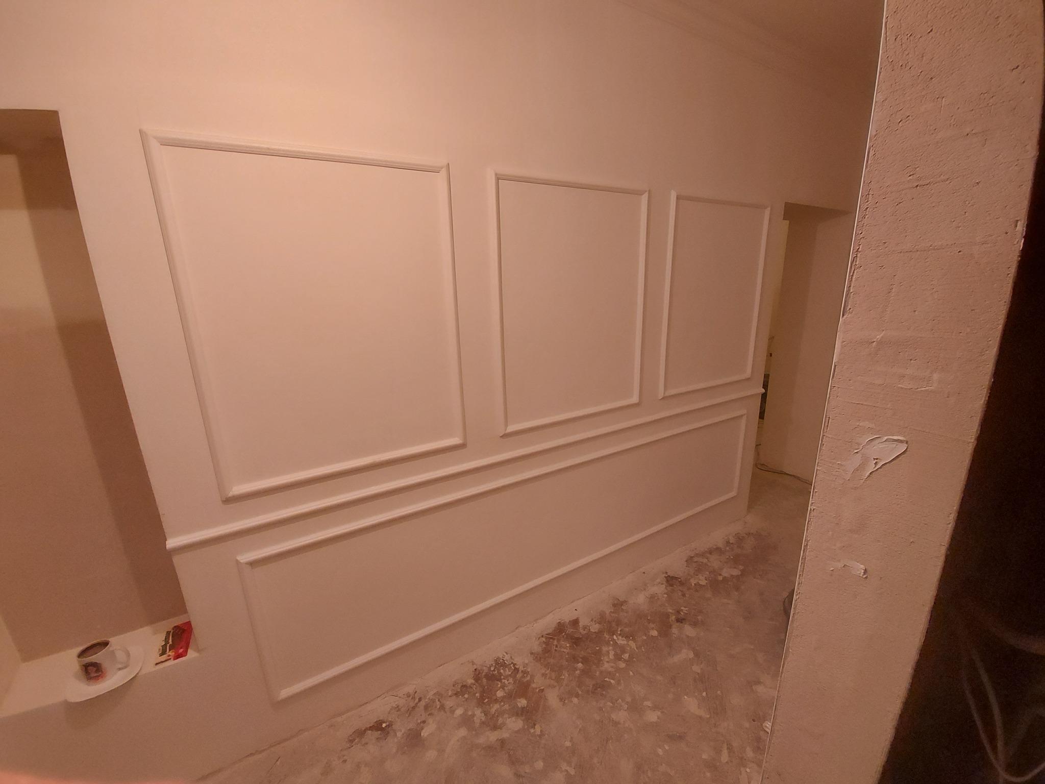 Ściana z zamontowanymi dekoracyjnymi panelami ściennymi w trakcie prac wykończeniowych, widoczna filiżanka kawy w niszy ściennej, surowa podłoga w tle.