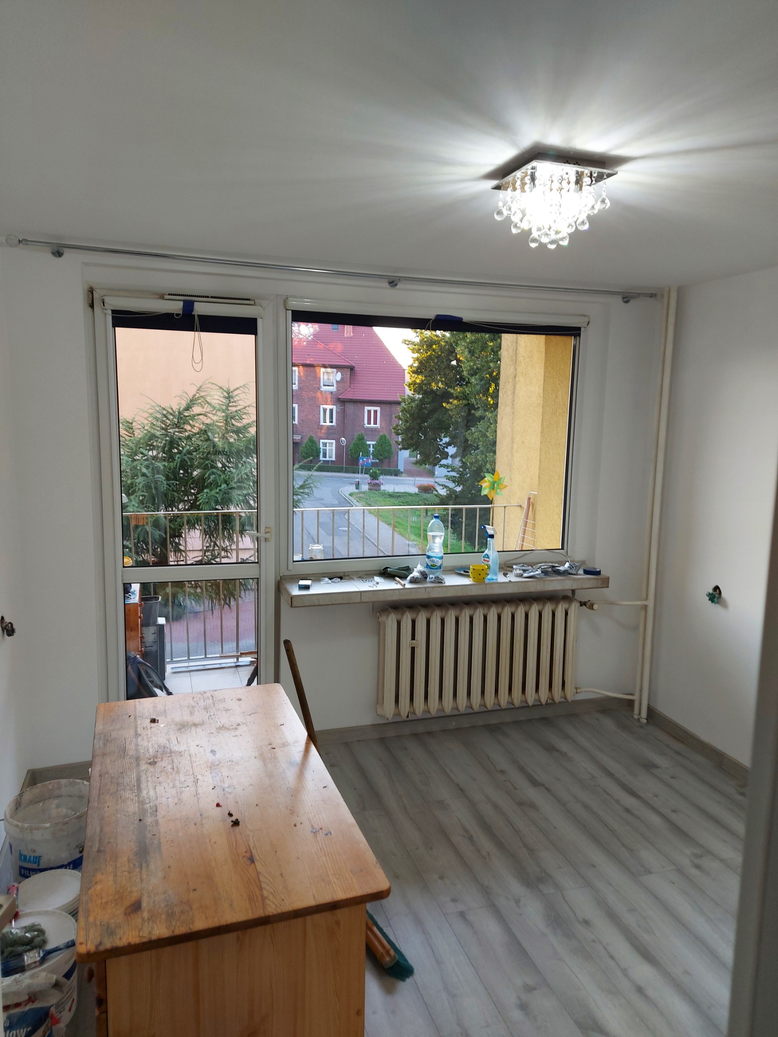 Pokój w trakcie odświeżania: surowe ściany, drewniany stół z farbami, grzejnik pod oknem z widokiem na zieleń, parkiet imitujący drewno i kryształowy plafon.
